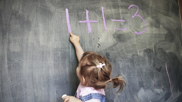 Two year old girl holding chalk pointing at a blackboard