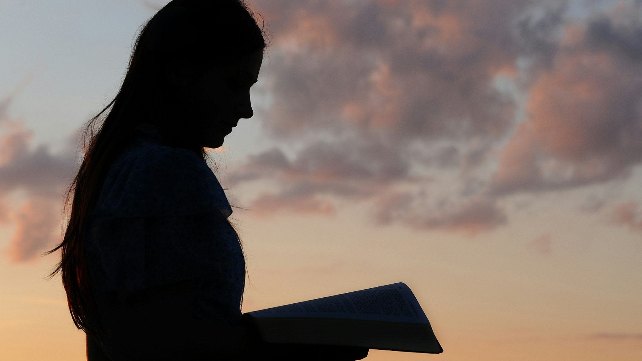 Silhouette of a young girl reading from Bible in sunset light.