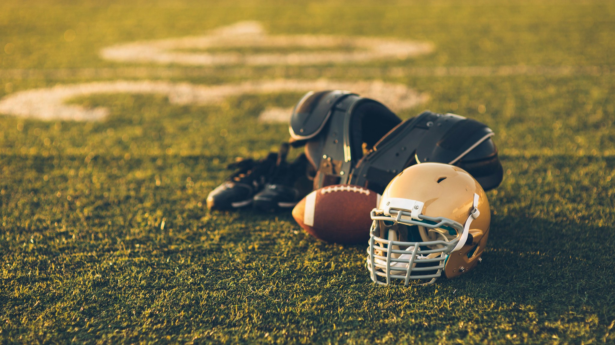A Gold American Football helmet sits with a football on a football playing field. The light is from the sun which is about to set, shallow depth of field. Copy space included. Sport background image.