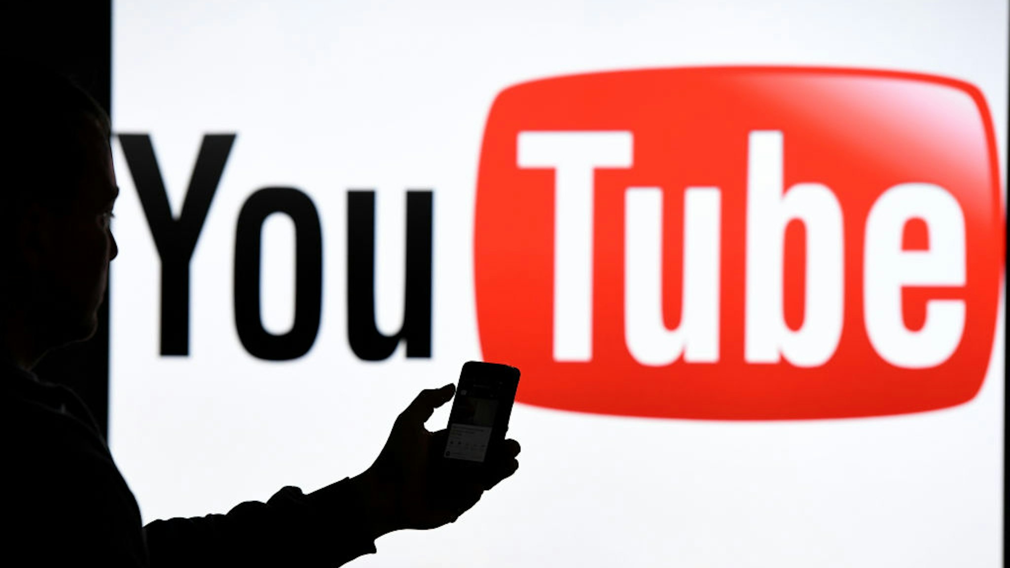 - A man with Smartphone stands before the Logo of the Internet platform YouTube. Photo: Carsten Rehder/dpa (Photo by Carsten Rehder/picture alliance via Getty Images)