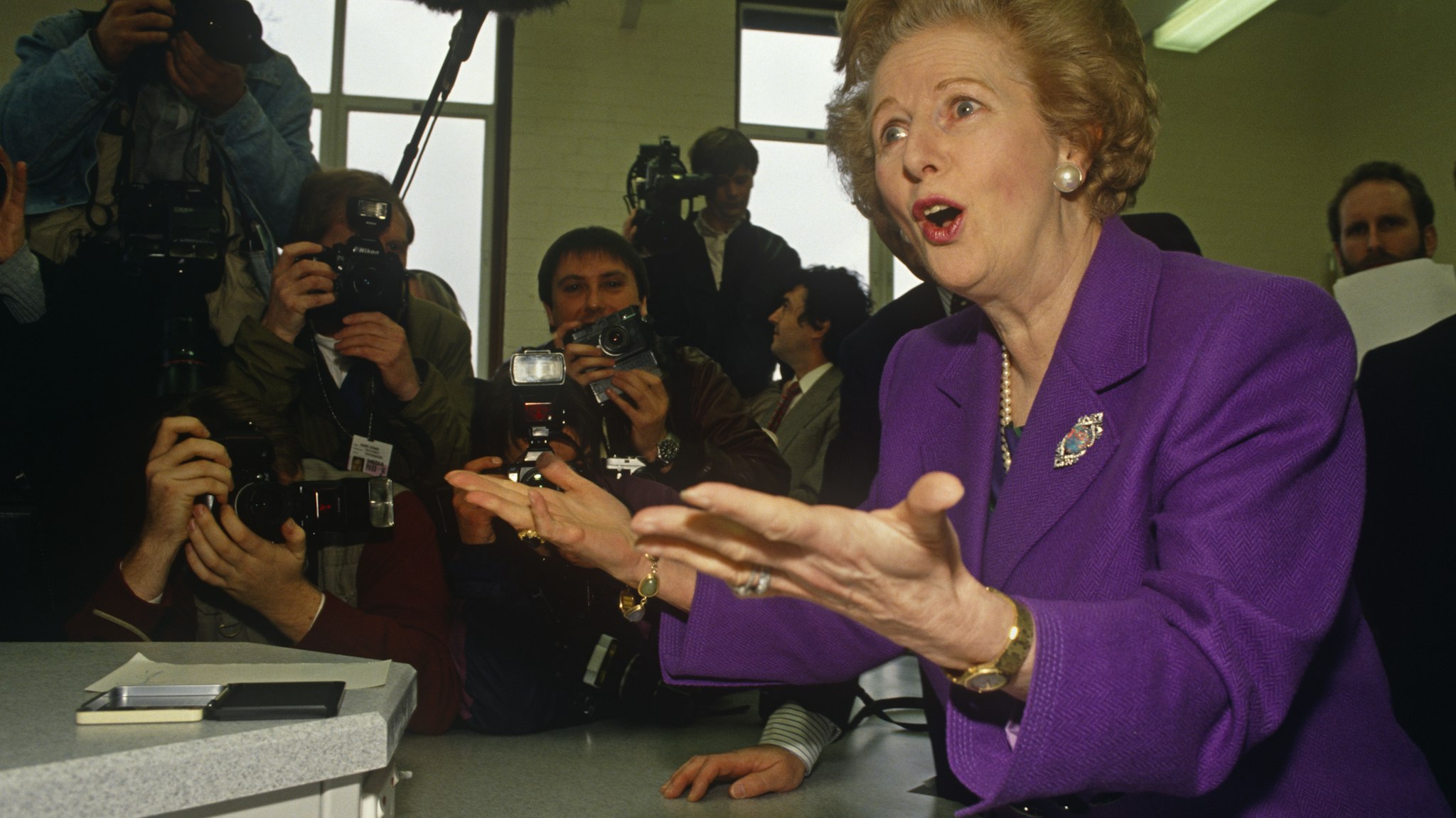 Margaret Thatcher plays up to the media at a North London school in her own constituency of Finchley during the 1992 general election. Although Thatcher had already resigned as Prime Minister in November 1990, John Major won the ensuing leadership election later that year. Photographers and cameramen surround the former-Prime Minister who is wearing a purple suit and matching broach. She is mid-sentence and has found something amusing to respond to the chants of the media. We see cameras, sound booms and flashes all prepared to photograph this famous statesman including Tom Stoddart who is making eye-contact with the viewer. (Photo by In Pictures Ltd./Corbis via Getty Images)