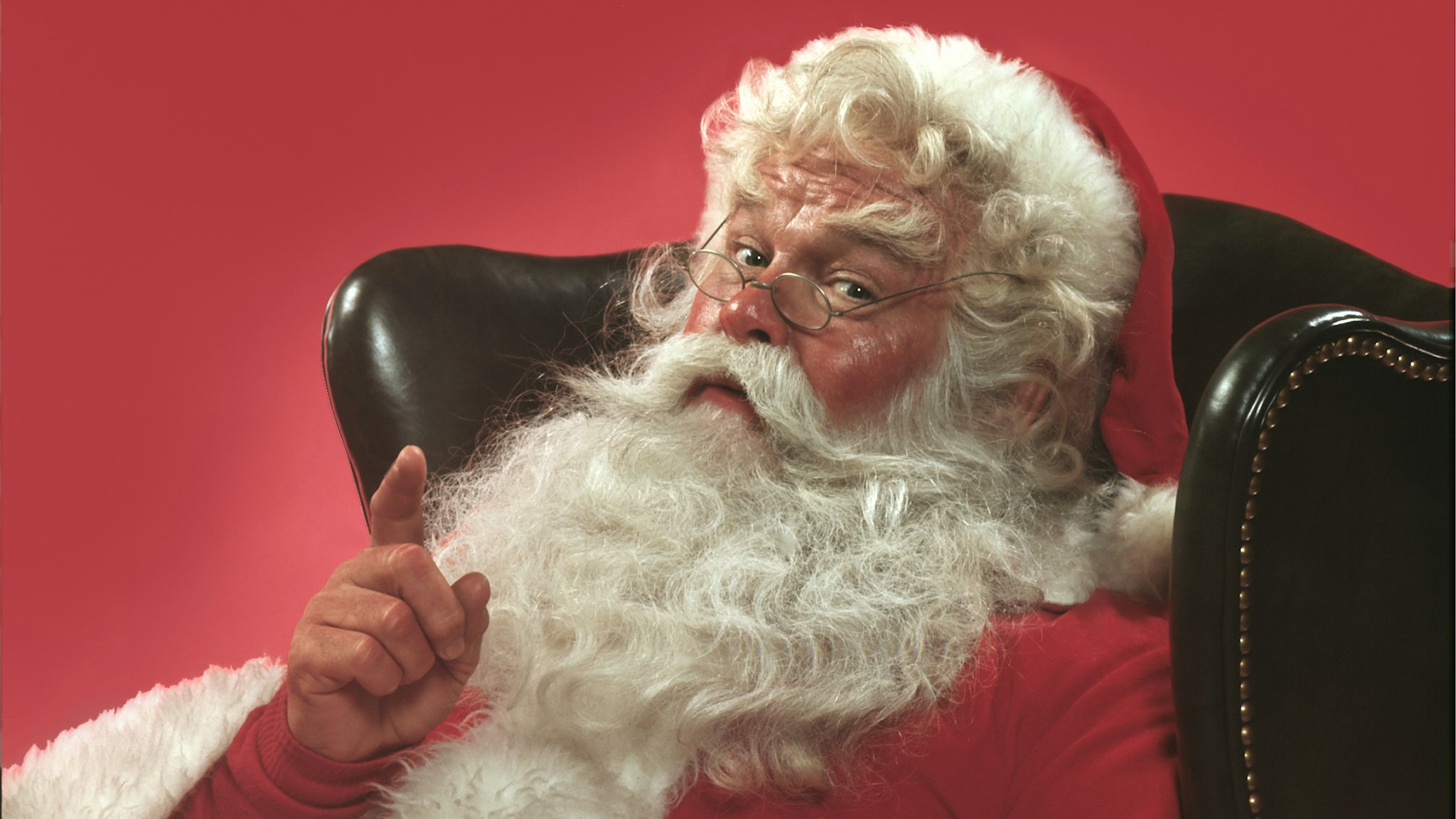 Portrait of Santa Claus sitting in a leather armchair raising one hand in a knowing gesture, 1980. United States.