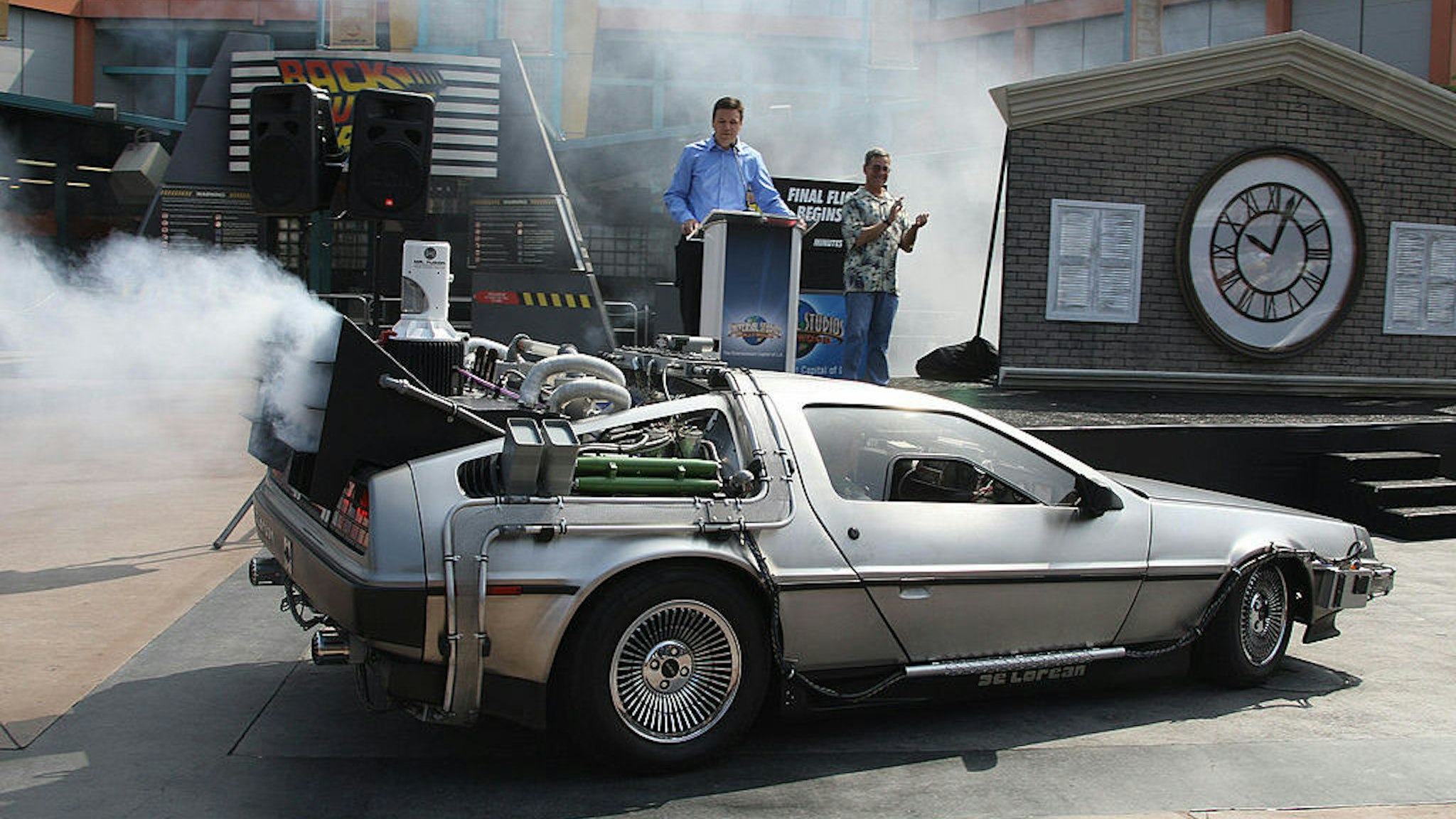 Actor Christopher Lloyd (in passenger seat) arrives in a DeLorean car at Universal Studios Hollywood's "Back to the Future - The Ride" in University City, California, 02 August 2007. Lloyd, who portrayed Doc Brown in the 1985 film "Back to the Future," made the apearence to mark a month-long countdown to the closure of the 14-year-old ride. The DeLorean Motor Co. (Texas), a suburban Houston company that rebuilds DeLoreans, recently announced plans to manufacture a limited number of new Deloreans in 2008. The last DeLorean rolled off the assembly line in Northern Ireland in 1982. On the podium are "Back to the Future" writer and co-creator Bob Gale (R) and Universal Studios Hollywood's Chris MacKenzie (L). AFP PHOTO / ROBYN BECK (Photo credit should read ROBYN BECK/AFP via Getty Images)