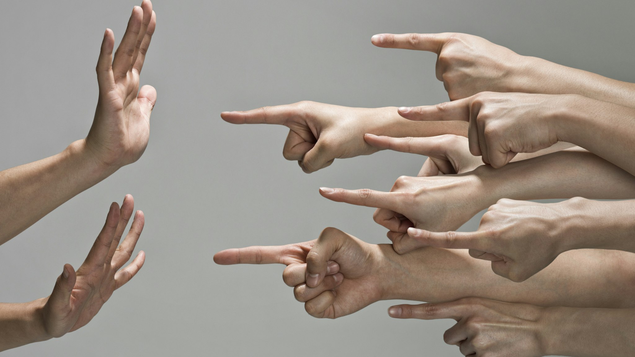 Multiple white hands are pointing index fingers from the right to the left at one set of hands. The pair of hands that are being pointed at are up in a defensive pose, with fingers to the sky and palms towards the pointing fingers.