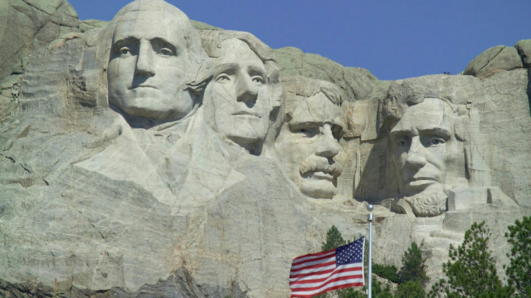 The faces of American Presidents George Washington, Thomas Jefferson, Theodore Roosevelt e Abraham Lincoln carved in the granite, American flag in the foreground, Mount Rushmore National Memorial, South Dakota, United States of America.
