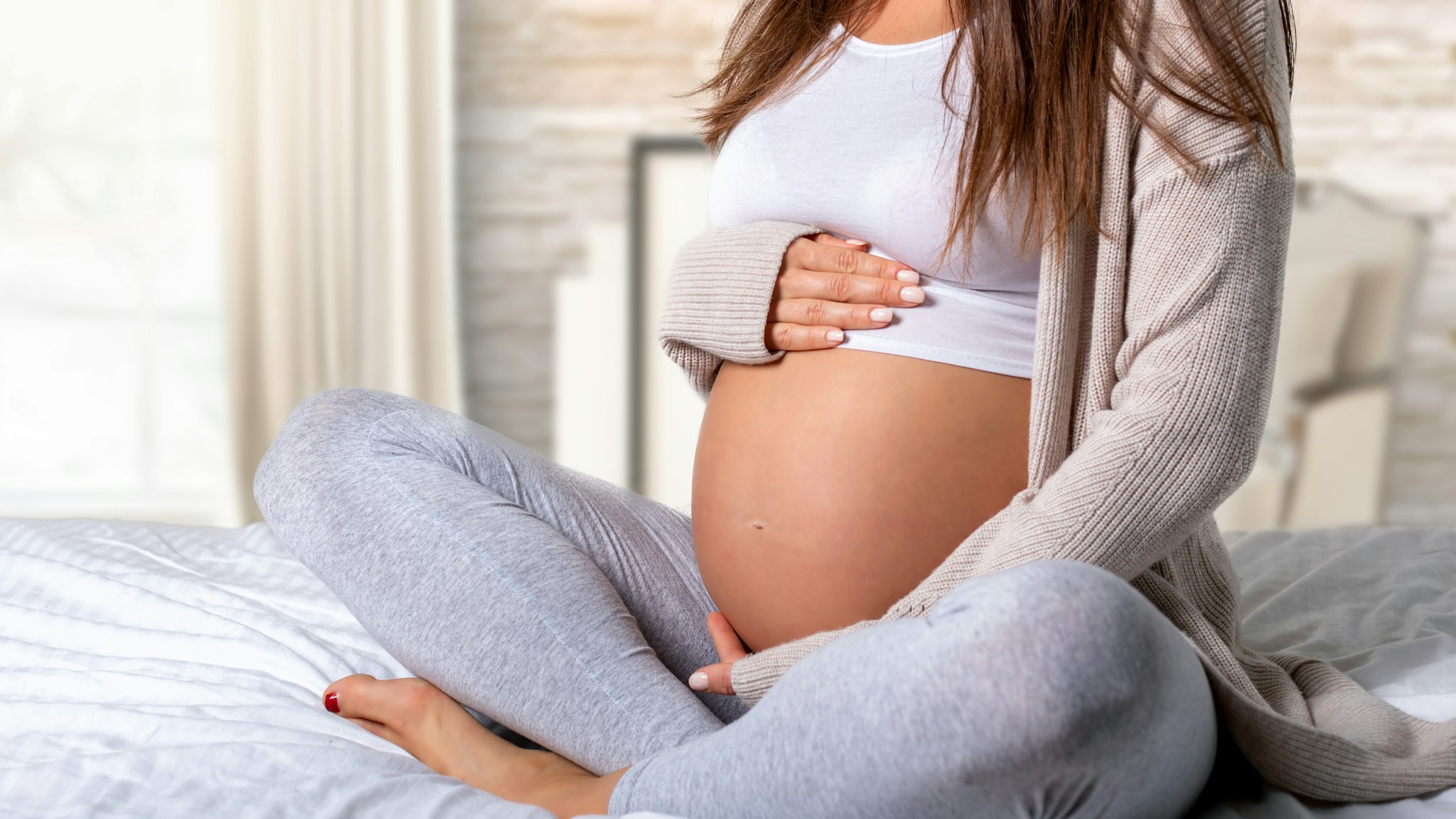 Low Section Of Pregnant Woman Touching Belly While Sitting On Bed - stock photo
