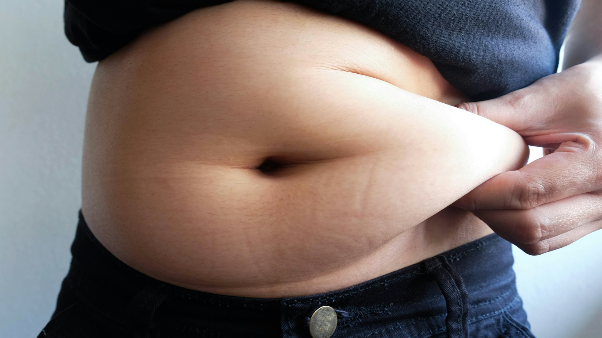 Midsection Of Overweight Woman Holding Stomach Fats While Standing Against Wall - stock photo