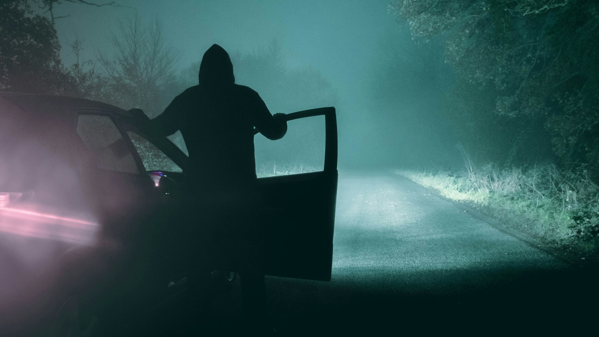 A lone, hooded figure standing next to a car looking at an empty misty winter country road silhouetted at night by car headlights - stock photo