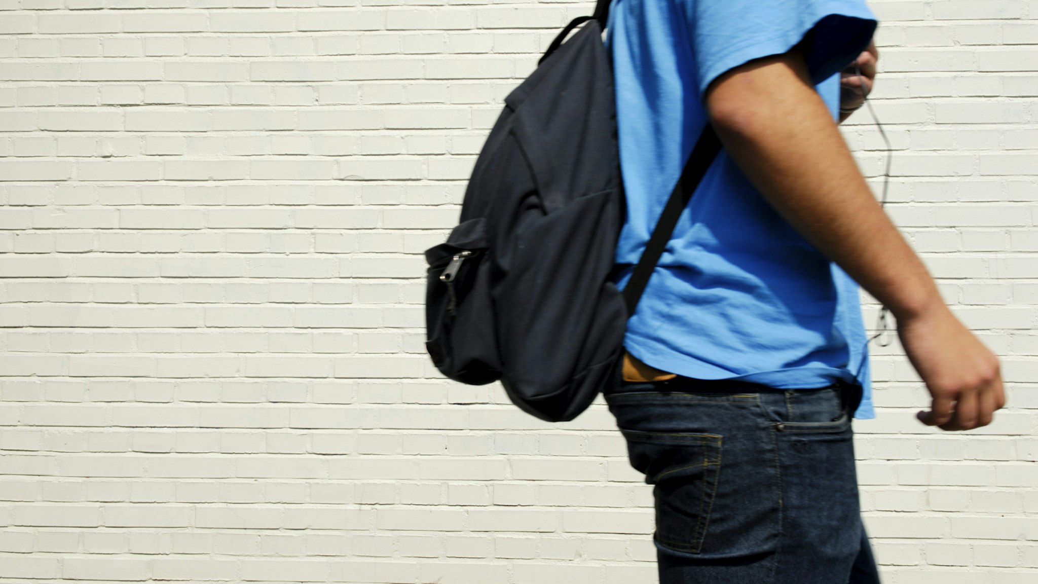 Young male student (with a black backpack, wearing a blue T-shirt and jeans) on his way to school passing an off-white brick wall. Focus is on the wall.