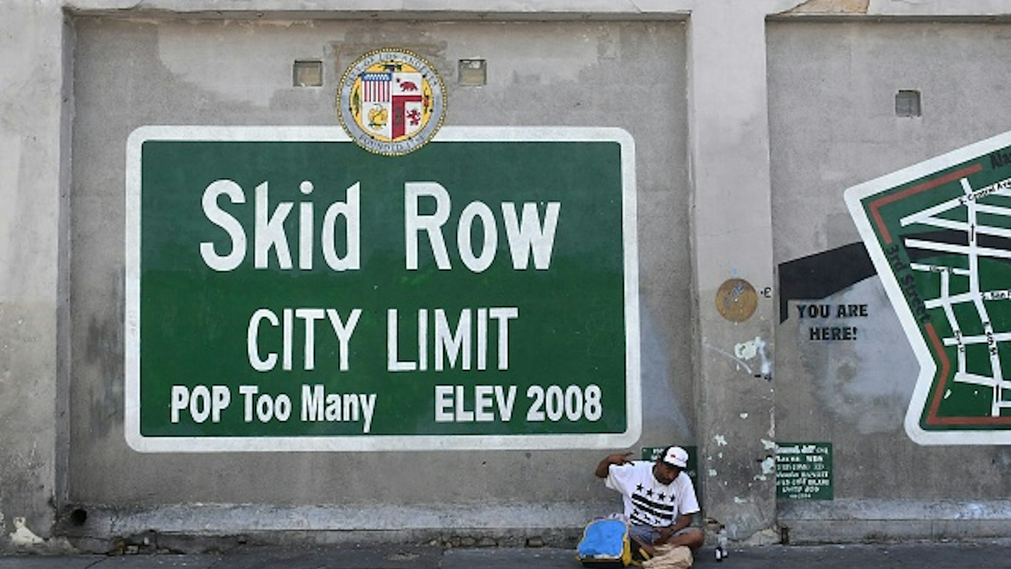 TOPSHOT - A man gestures while seated beside a Skid Row painting on a sidewalk in downtown Los Angeles on May 30, 2019. - The city of Los Angeles on May 29 agreed to allow homeless people on Skid Row to keep their property and not have it seized, providing the items are not bulky or hazardous.