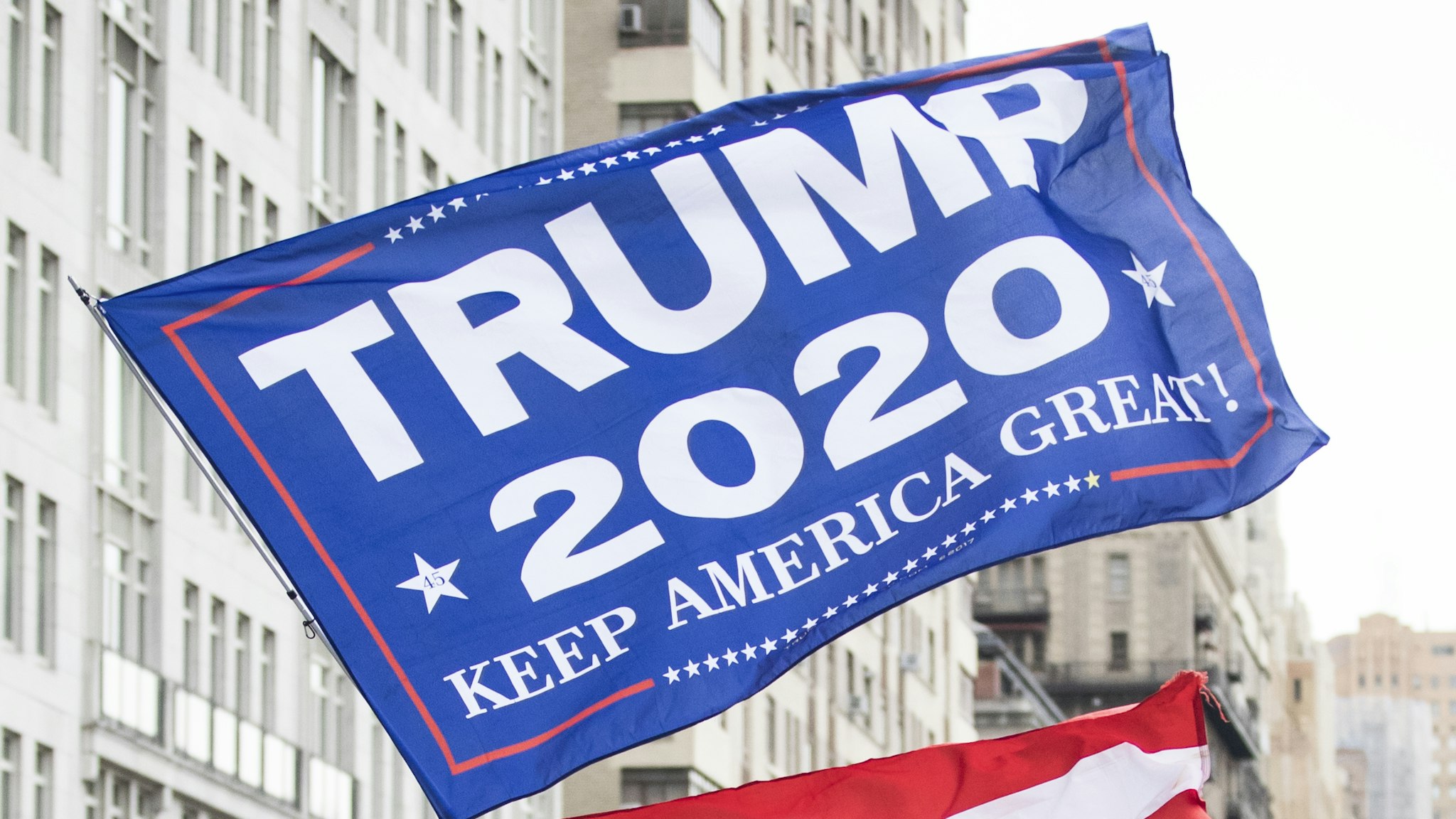 MANHATTAN, NY - JANUARY 18: A Trump supporter waves a huge flag with TRUMP 2020 Keep America Great! walks in front of the marchers as they start the march on Central Park West during the Woman's March in the borough of Manhattan in NY on January 18, 2020, USA. The rally took place 3 years after the inauguration of President Donald Trump and 3 days after the Articles of Impeachment were brought to the Senate. Thousands gather to protest equal rights at the 2020 Women's March.