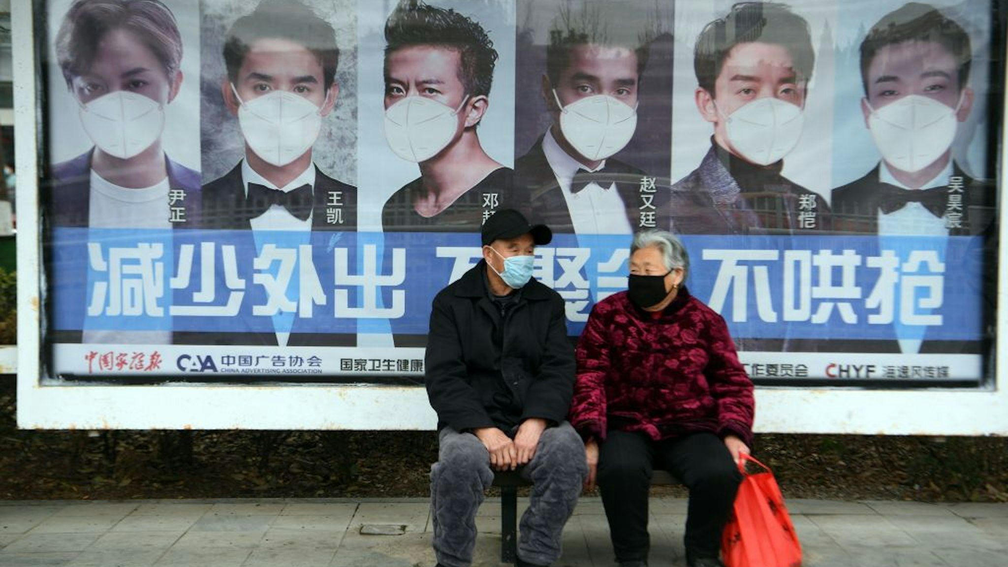 A man and woman sit in front of a poster reminding citizens to wear face masks as a preventive measure against the COVID-19 coronavirus, at a bus stop in Bozhou, in China's eastern Anhui province on March 6, 2020. - China reported 30 more deaths from the new coronavirus outbreak on March 6, with fresh infections rising for a second straight day and 16 new cases imported from overseas. (Photo by STR / AFP)