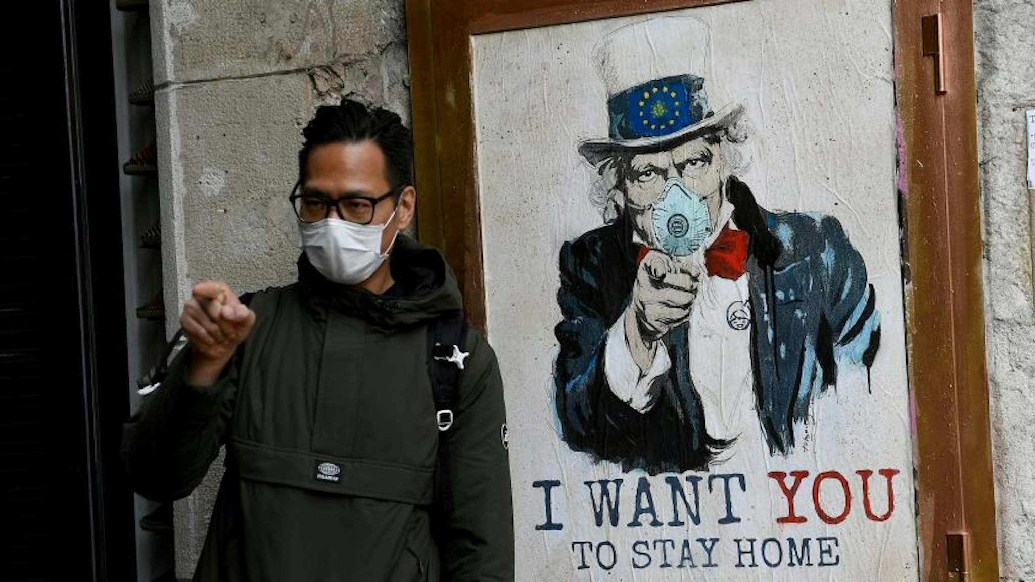 A man wearing a face mask poses with a poster by artist TVBoy featuring Uncle Sam and reading "I want you to stay home" in Barcelona on March 14, 2020 after regional authorities ordered all shops in the region be shuttered from today through March 26, save for those selling food, chemists and petrol stations, in order to slow the coronavirus spread. (Photo by Josep LAGO / AFP) / RESTRICTED TO EDITORIAL USE - MANDATORY MENTION OF THE ARTIST UPON PUBLICATION - TO ILLUSTRATE THE EVENT AS SPECIFIED IN THE CAPTION (Photo by