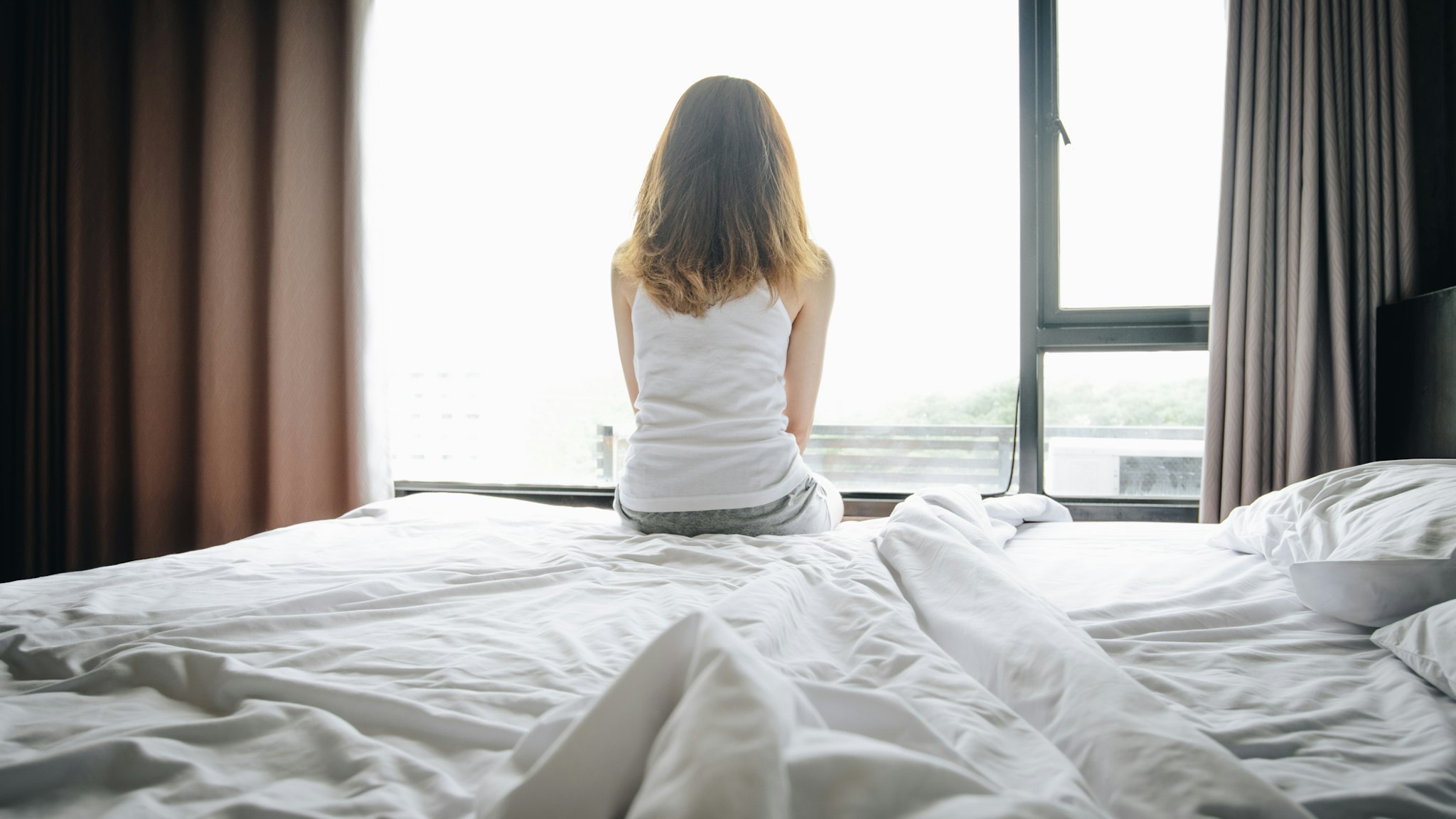 Portrait of depressed woman sitting alone on bed, looking to outside the window. - stock photo