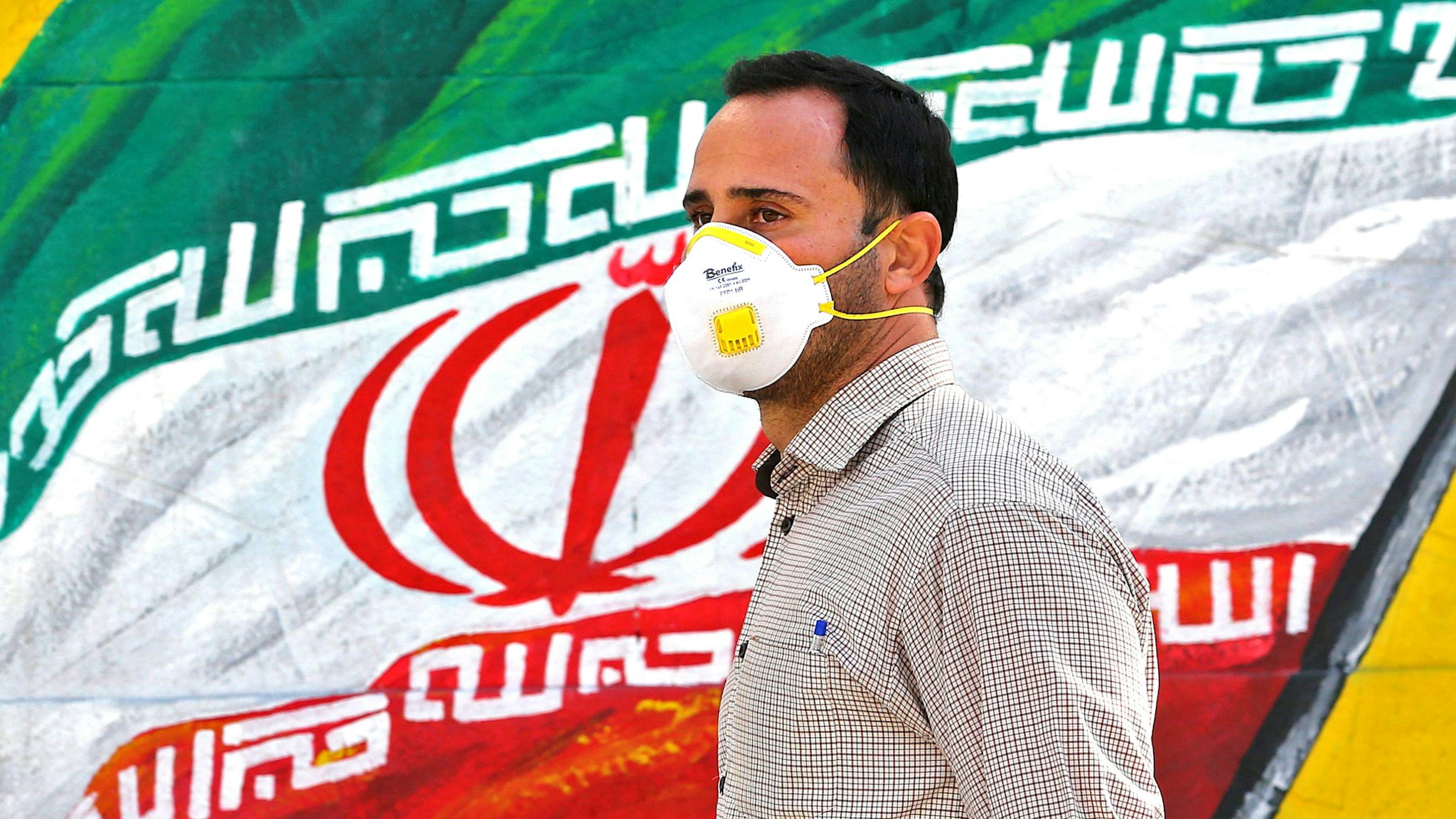An Iranian man wearing a mask walks past a mural displaying his national flag in Tehran on March 4, 2020. - Iran has scrambled to halt the rapid spread of the COVID-19 virus, shutting schools and universities, suspending major cultural and sporting events, and cutting back on work hours.