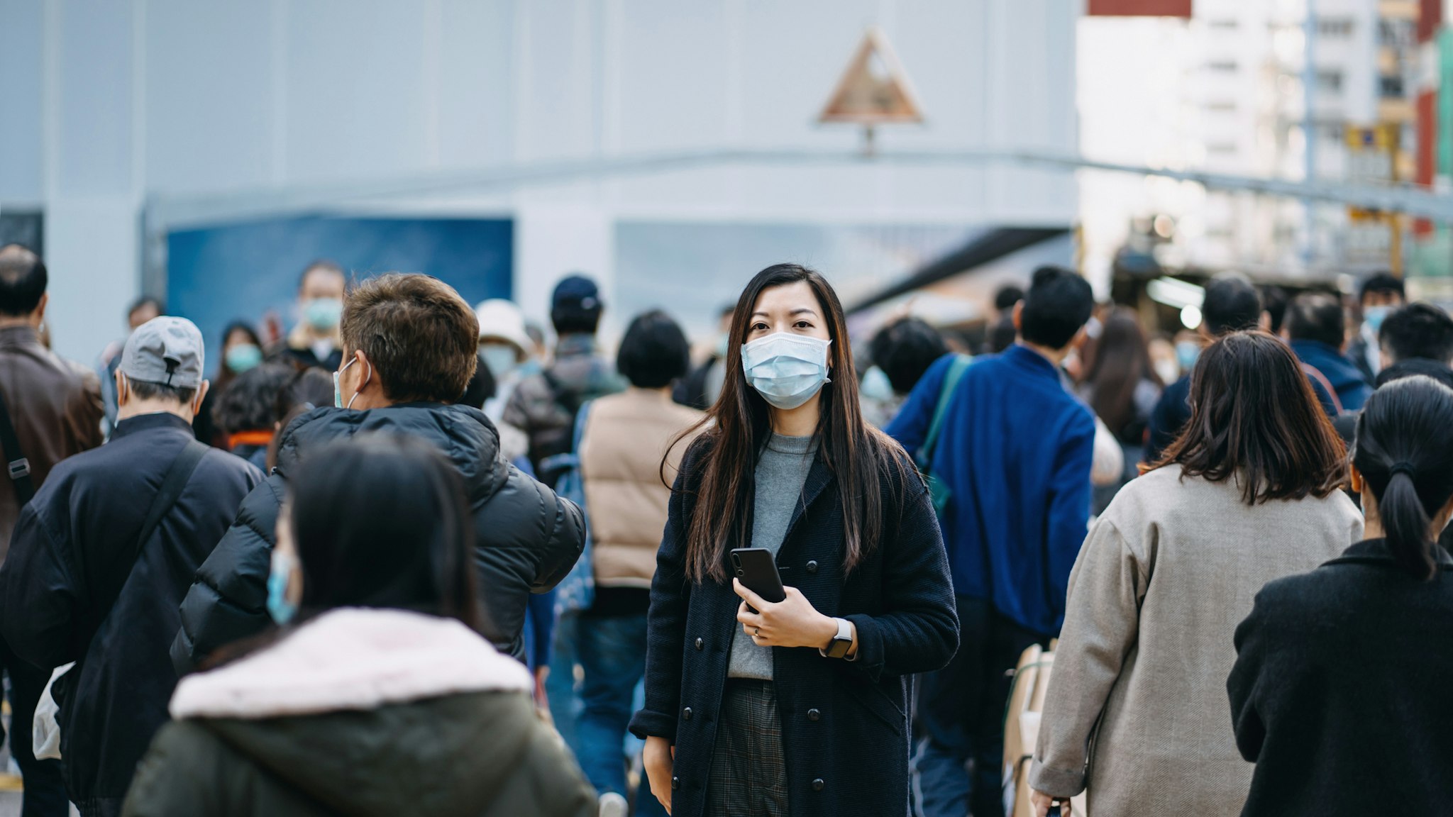 Asian woman with protective face mask holding smartphone standing in the middle of busy downtown city street amidst crowd of pedestrians passing by