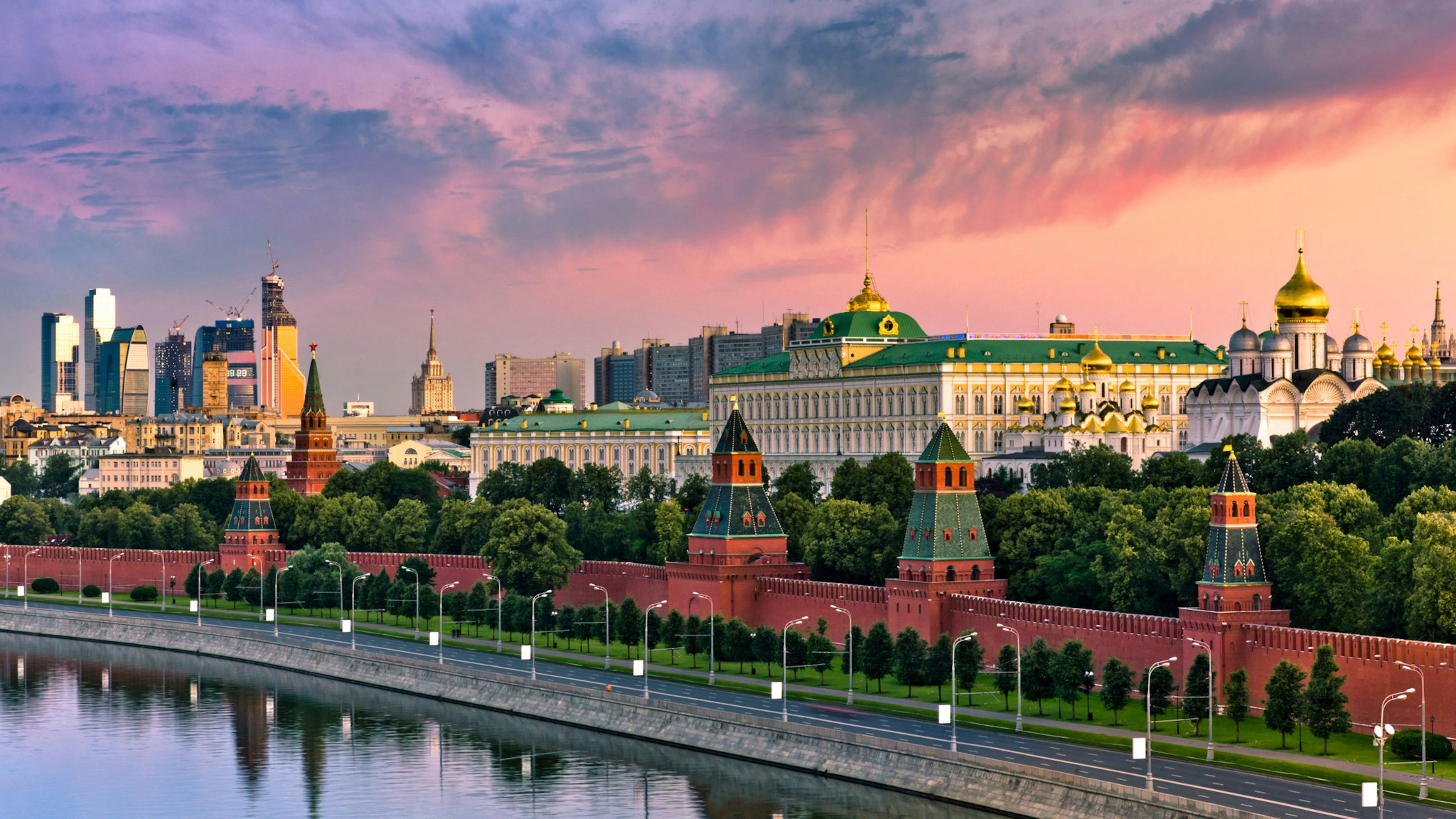 View of the Moskva River along the Kremlin wall in Moscow, Russia, with water and a walkway in the foreground and a beautiful sunset sky in the background. The image shows the Grand Kremlin Palace and the cathedrals of the Moscow Kremlin. The view is from the top of the Hotel Baltschug Kempinski.