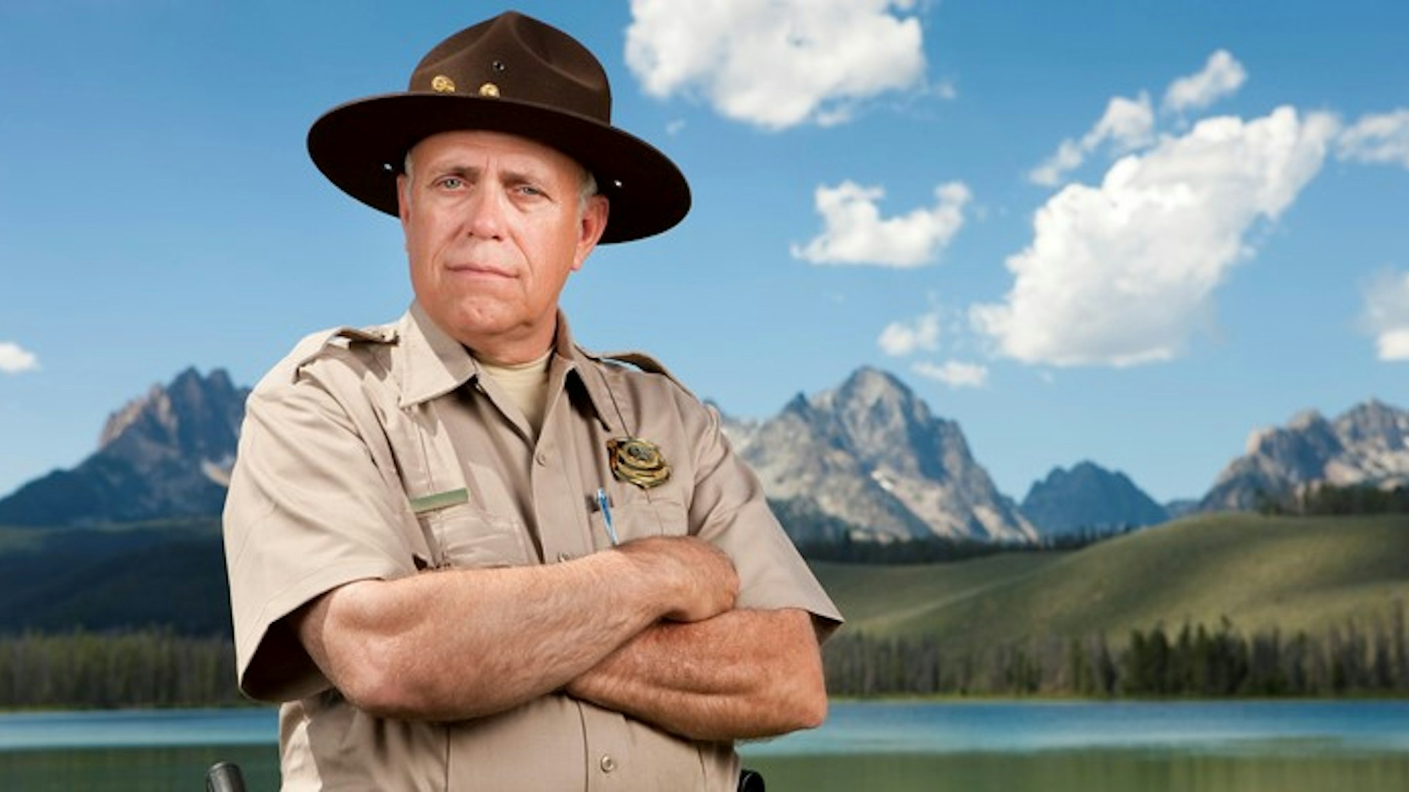A mature male park ranger (or police officer) standing in front of a beautiful scenic backdrop of tall mountains and a mirror lake.