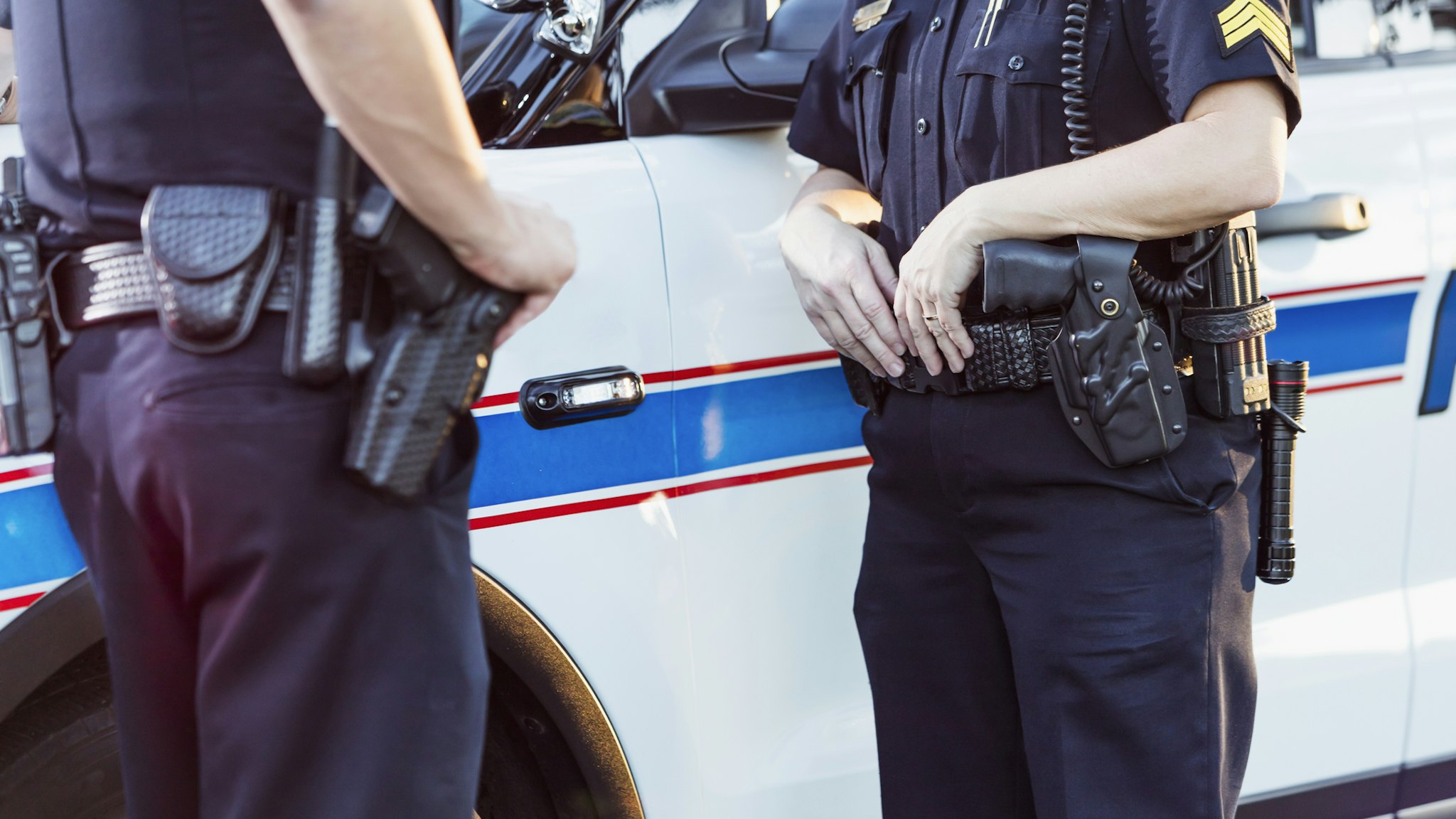 Cropped view of police officers standing beside a squad car having a conversation. The focus is on the policewoman whose hands are resting on her gun belt.