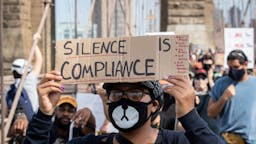 Silence Is Compliance A protester wearing a mask that resembles a cat holds a handmade sign that says,"Silence is Compliance" with the cables of the Brooklyn Bridge behind them as they walk across the Brooklyn Bridge in protest of police racism. Protesters walked from Brooklyn to Manhattan after a memorial ceremony in Brooklyn with Terrence Floyd, brother of George Floyd. Protesters have taken to the streets across America and around the world after the killing of George Floyd at the hands of a white police officer Derek Chauvin that was kneeling on his neck during his arrest as he pleaded that he couldn't breathe. The protest are attempting to give a voice to the need for human rights for African American's and to stop police brutality against people of color. Many people were wearing masks and observing social distancing due to the coronavirus pandemic. Terrence Floyd echoed the word of peaceful protest saying that violent protests are overshadowing What's Going On. New York City remains in a curfew as an answer to nights of unrest looting and destruction. Photographed in the Manhattan Borough of New York on June 04, 2020, USA. (Photo by Ira L. Black/Corbis via Getty Images)