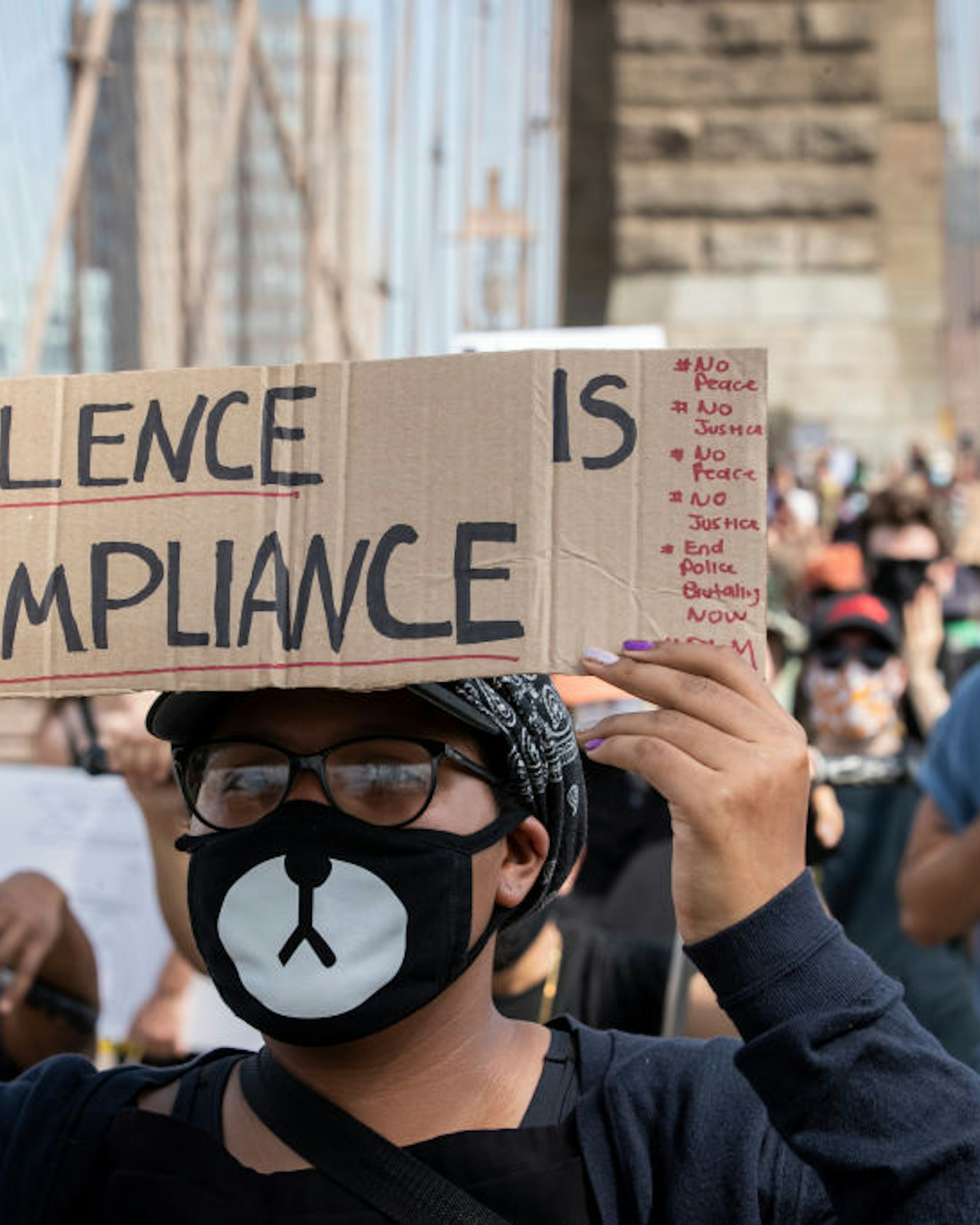 A protester wearing a mask that resembles a cat holds a handmade sign that says,"Silence is Compliance" with the cables of the Brooklyn Bridge behind them as they walk across the Brooklyn Bridge in protest of police racism. Protesters walked from Brooklyn to Manhattan after a memorial ceremony in Brooklyn with Terrence Floyd, brother of George Floyd. Protesters have taken to the streets across America and around the world after the killing of George Floyd at the hands of a white police officer Derek Chauvin that was kneeling on his neck during his arrest as he pleaded that he couldn't breathe. The protest are attempting to give a voice to the need for human rights for African American's and to stop police brutality against people of color. Many people were wearing masks and observing social distancing due to the coronavirus pandemic. Terrence Floyd echoed the word of peaceful protest saying that violent protests are overshadowing What's Going On. New York City remains in a curfew as an answer to nights of unrest looting and destruction. Photographed in the Manhattan Borough of New York on June 04, 2020, USA. (Photo by Ira L. Black/Corbis via Getty Images)