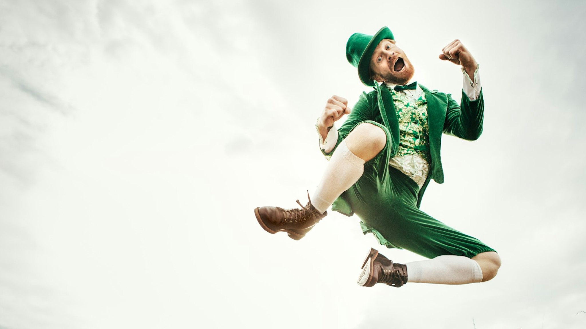 A stereotypical Irish character all ready for Saint Patricks day jumps and dances in an open field of Irish country side. Copy space in the sky and grass.