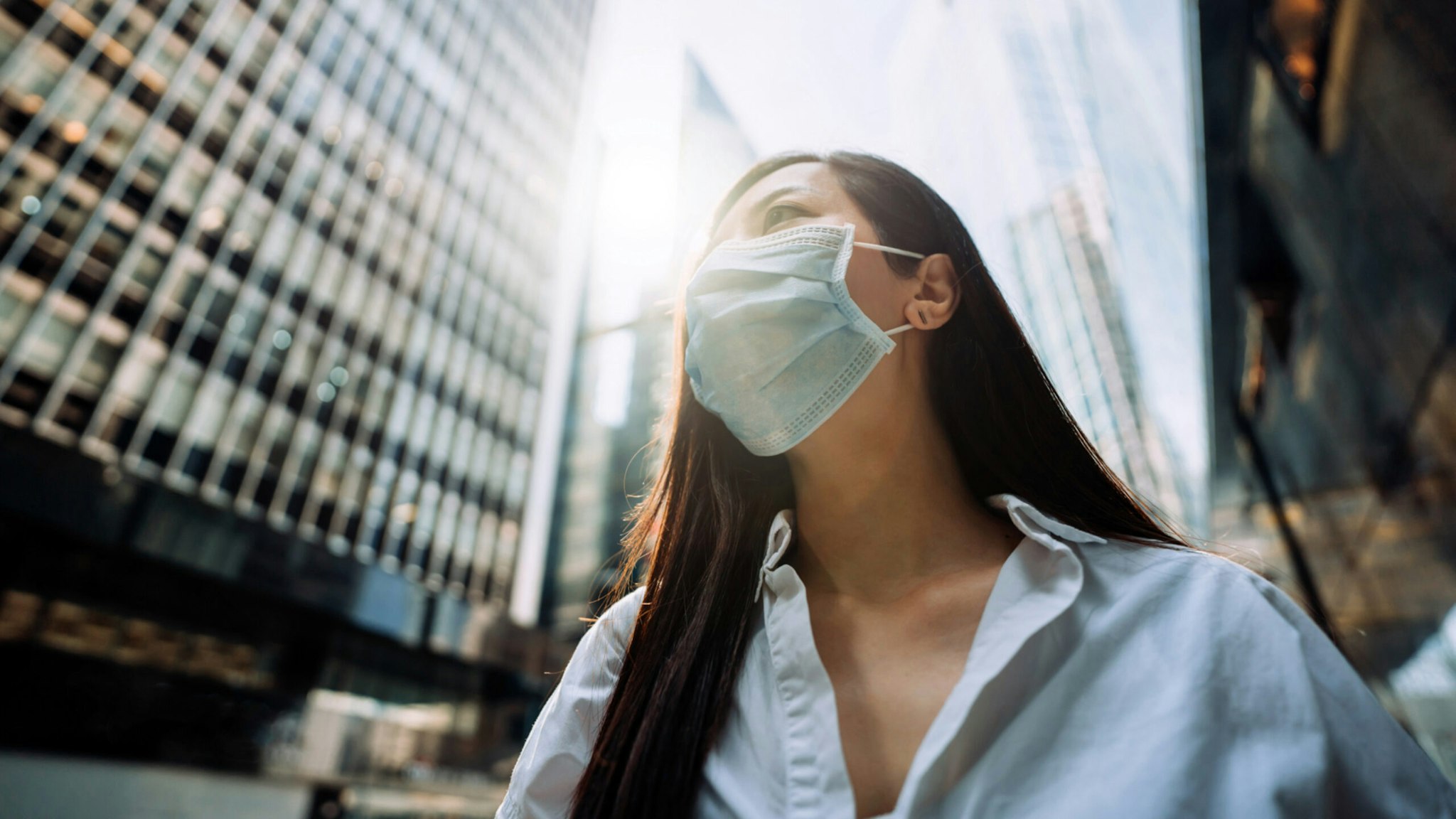 Young Asian businesswoman with protective face mask to protect and prevent from the spread of viruses during the coronavirus health crisis, standing in an energetic and prosperous downtown city street against corporate skyscrapers.