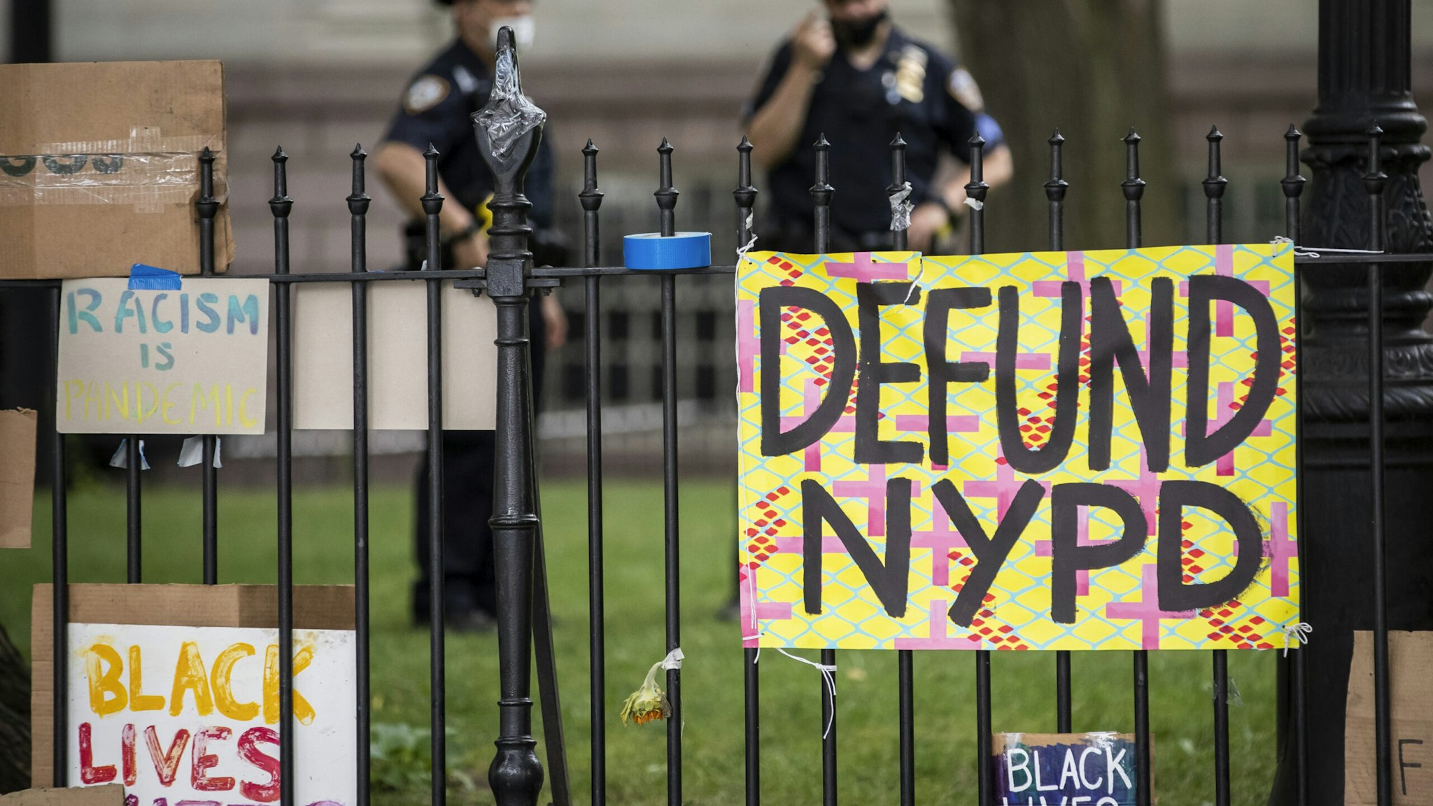 MANHATTAN, NY - JUNE 30: Protesters signs hang on the gate in City Hall Park during the Occupy City Hall protests that say, "Defund NYPD" and "BLM" with NYPD police officers in the background. Earlier in the day, one of the members of Warriors in the Garden was beaten and arrested by police. This was part of the Occupy City Hall effort which has seen protesters camping out for days until today where New York City Council is to vote on the NYPD Budget. Protesters continue taking to the streets across America and around the world after the killing of George Floyd at the hands of a white police officer Derek Chauvin that was kneeling on his neck during for eight minutes, was caught on video and went viral. During his arrest as Floyd pleaded, "I Can't Breathe". The protest are attempting to give a voice to the need for human rights for African American's and to stop police brutality against people of color. They are also protesting deep-seated racism in America. Many people were wearing masks and observing social distancing due to the coronavirus pandemic. Photographed in the Manhattan Borough of New York on June 30, 2020, USA. (Photo by Ira L. Black/Corbis via Getty Images)