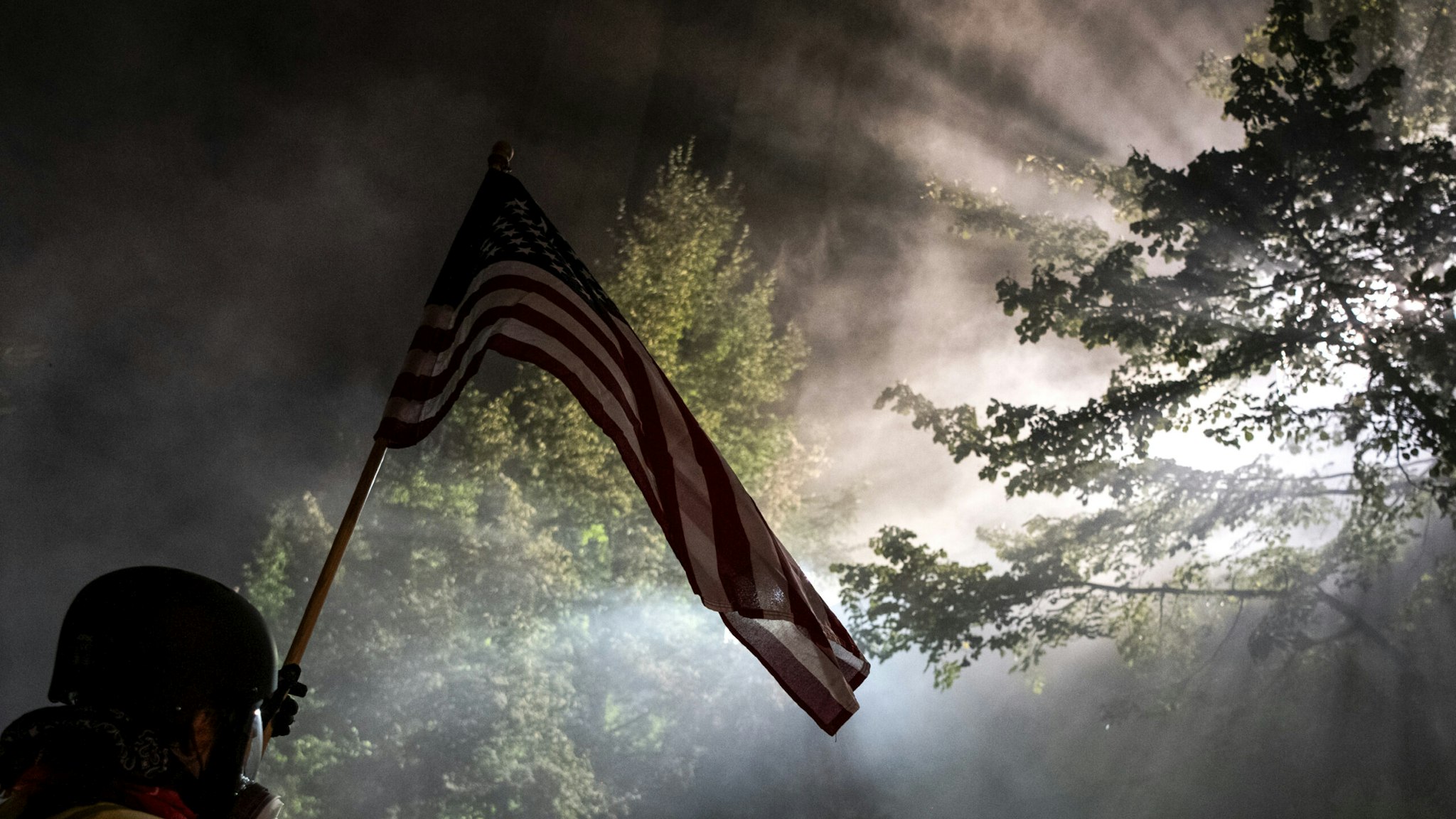 A demonstrators holding a US flag stands in a cloud of tear gas outside the Mark O. Hatfield Federal Courthouse during a night of protest against racial injustice, police brutality and the deployment of federal troops to US cities on July 29, 2020 in Portland, Oregon. - Protests in the US city of Portland have continued for more than 60 days. President Donald Trump's administration on July 29 agreed to a deal to defuse weeks of clashes with the withdrawal of federal forces whose presence enraged protesters, but the timing remained in dispute. (Photo by Alisha JUCEVIC / AFP) (Photo by ALISHA JUCEVIC/AFP via Getty Images)