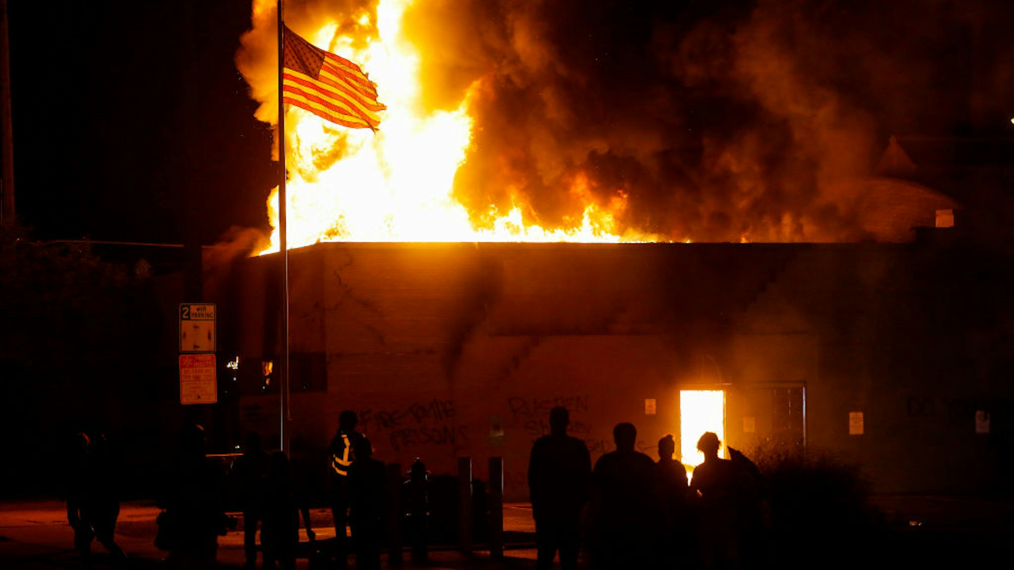 KENOSHA, WI - AUGUST 24: People watch a the American flag flies over a burning building during a riot as demonstrators protest the police shooting of Jacob Blake on Monday, August 24, 2020 in Kenosha, Wisconsin. Blake was shot in the back multiple times by police officers responding to a domestic dispute call yesterday.
