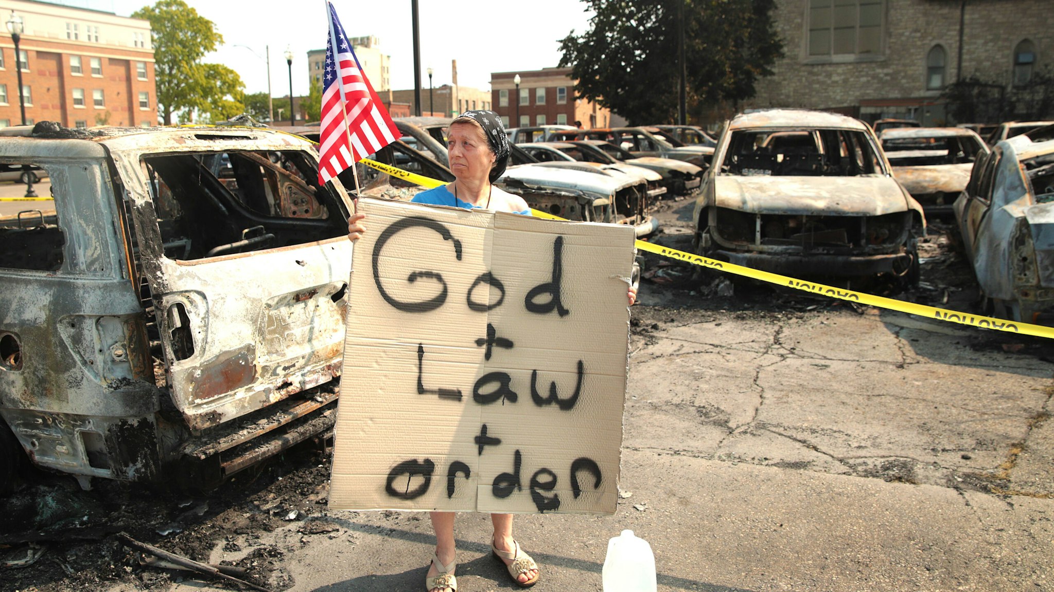 KENOSHA, WISCONSIN - AUGUST 24: A women holds a sign while standing in a used car lot with burned out cars after a night of unrest, on August 24, 2020 in Kenosha, Wisconsin. The unrest stemmed from an incident in which police shot a Black man multiple times in the back as he entered the driver's side door of a vehicle.