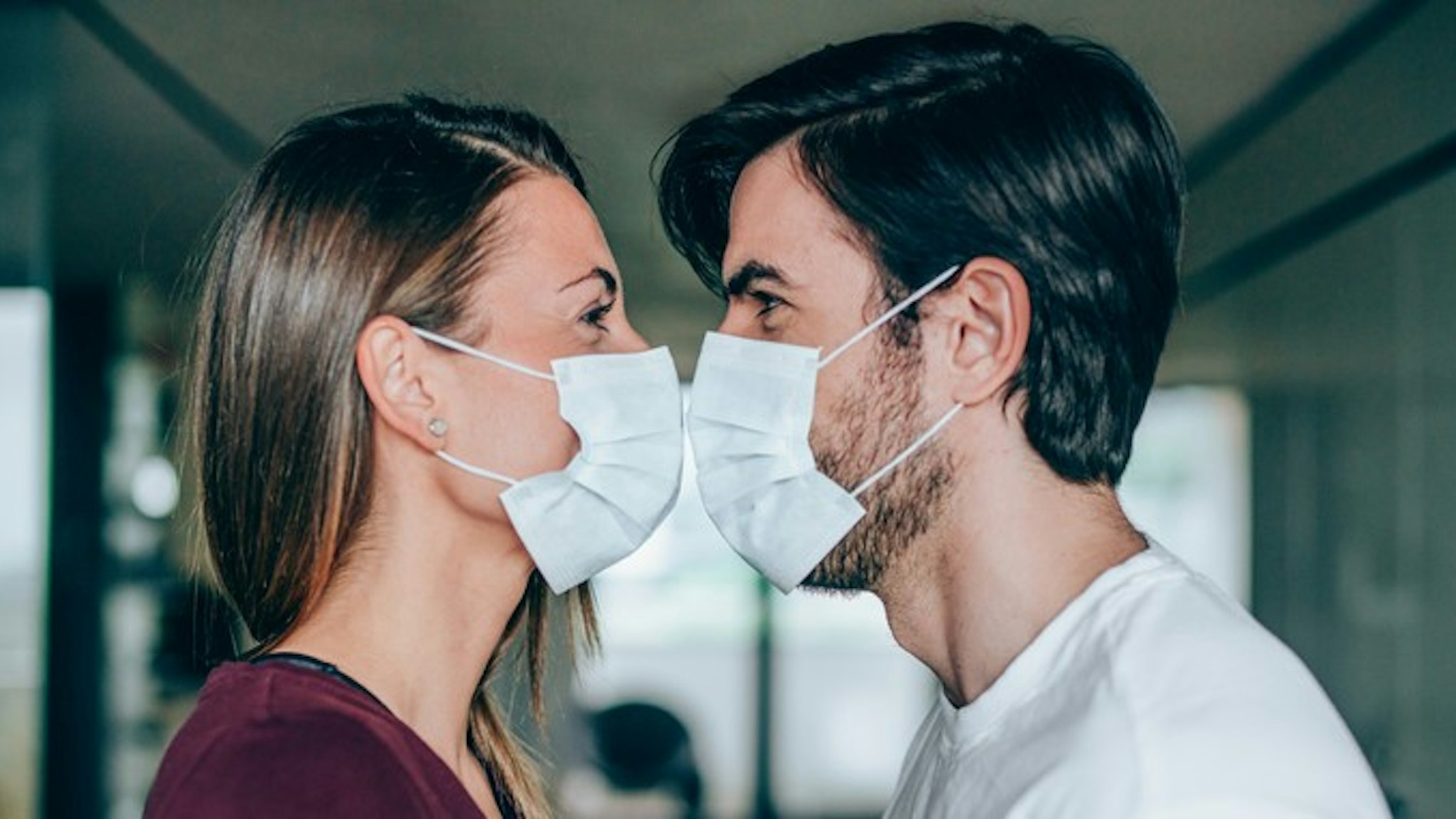 Beautiful happy young couple wearing protective face masks and kissing each other. Side view of young man and young woman in medical masks kissing while they are in home isolation during coronavirus/COVID-19 quarantine.