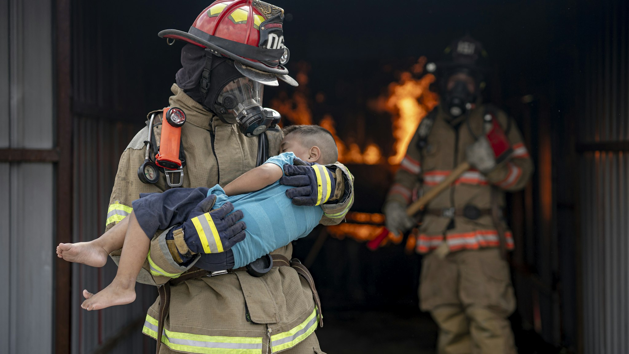 Brave Fireman of a Burning Building and Holds Saved boy in His Arms. Open fire and one Firefighter in the Background.