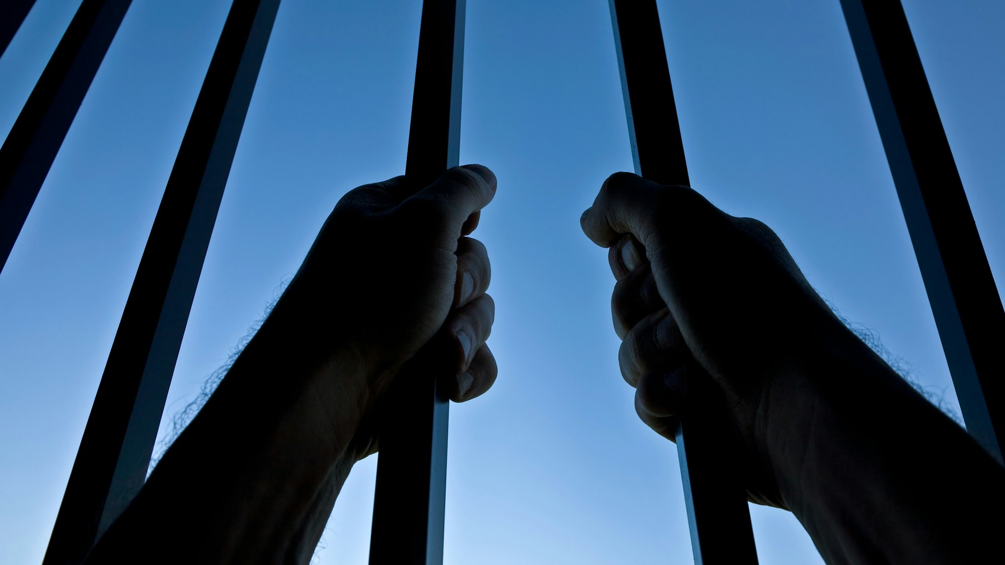 Silhouette of Hands Behind Jail Bars Against Clear Blue Sky - stock photo
