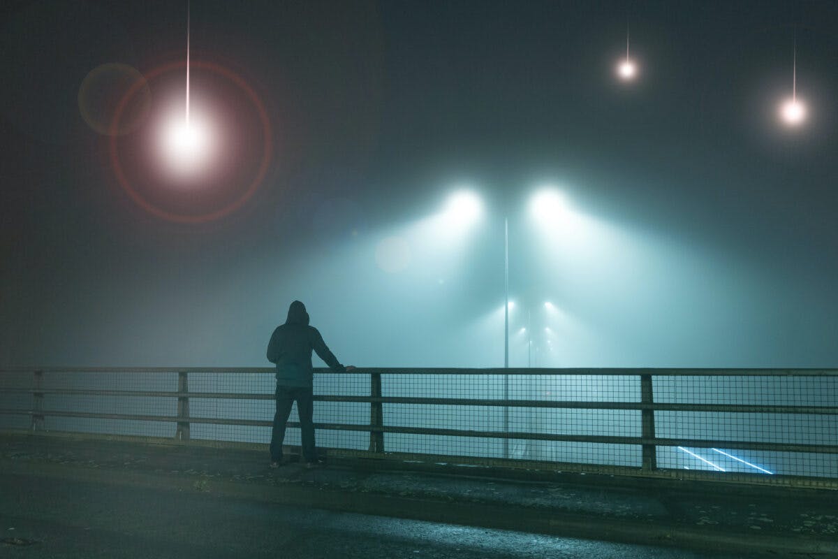 A hooded figure, standing with back to camera on a bridge, looking at UFO alien spaceships coming downfrom the sk., Street lights. On a foggy night.