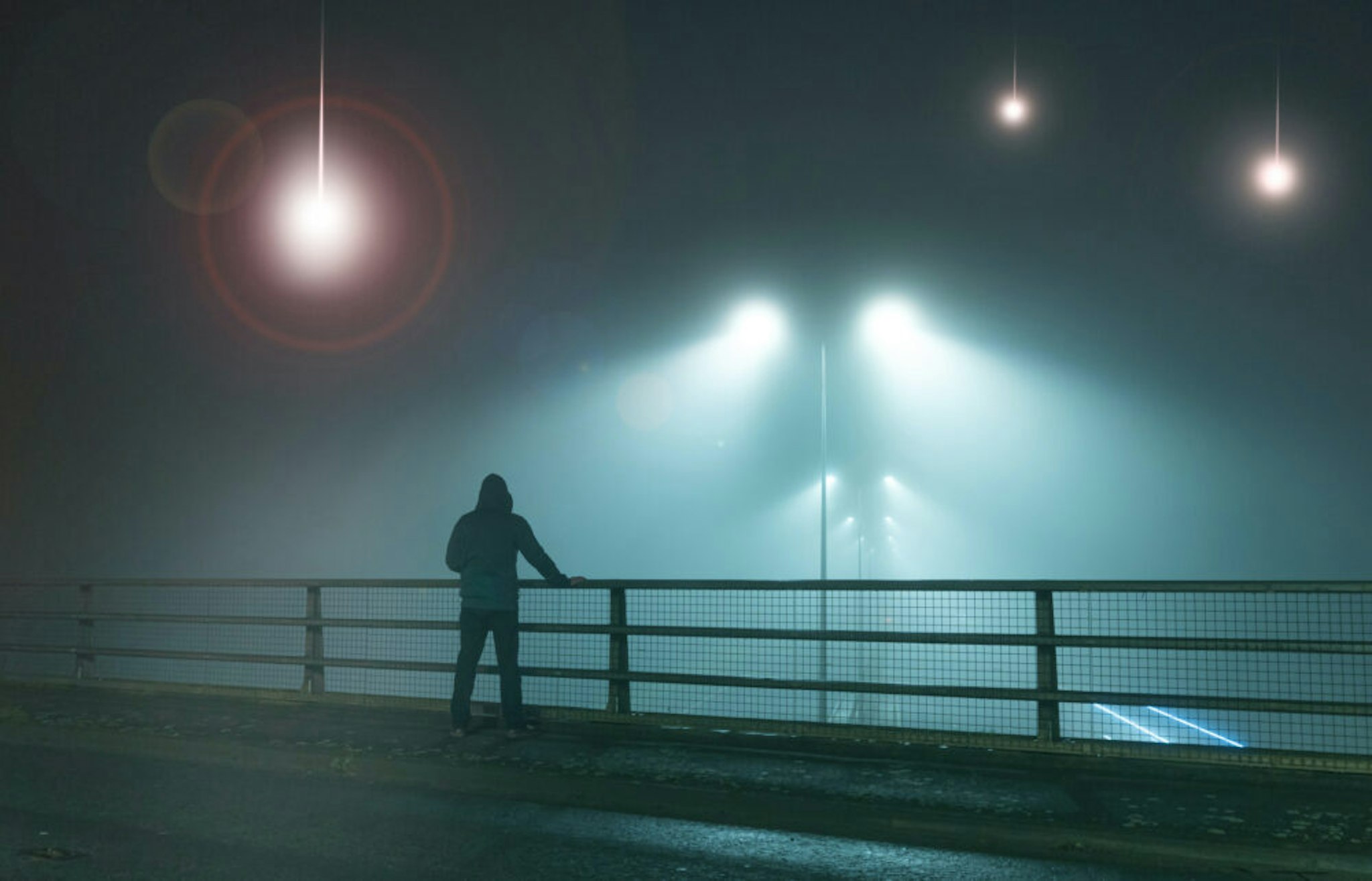 A hooded figure, standing with back to camera on a bridge, looking at UFO alien spaceships coming downfrom the sk., Street lights. On a foggy night.