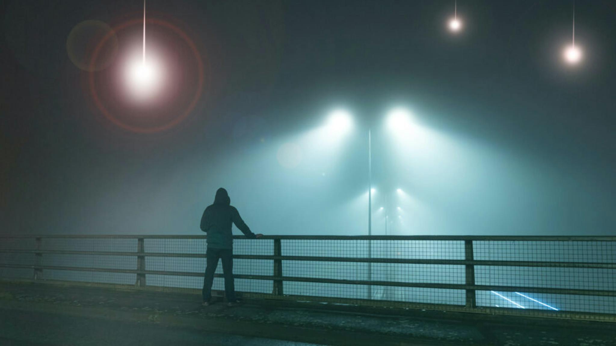 UFO A hooded figure, standing with back to camera on a bridge, looking at UFO alien spaceships coming downfrom the sk., Street lights. On a foggy night.
