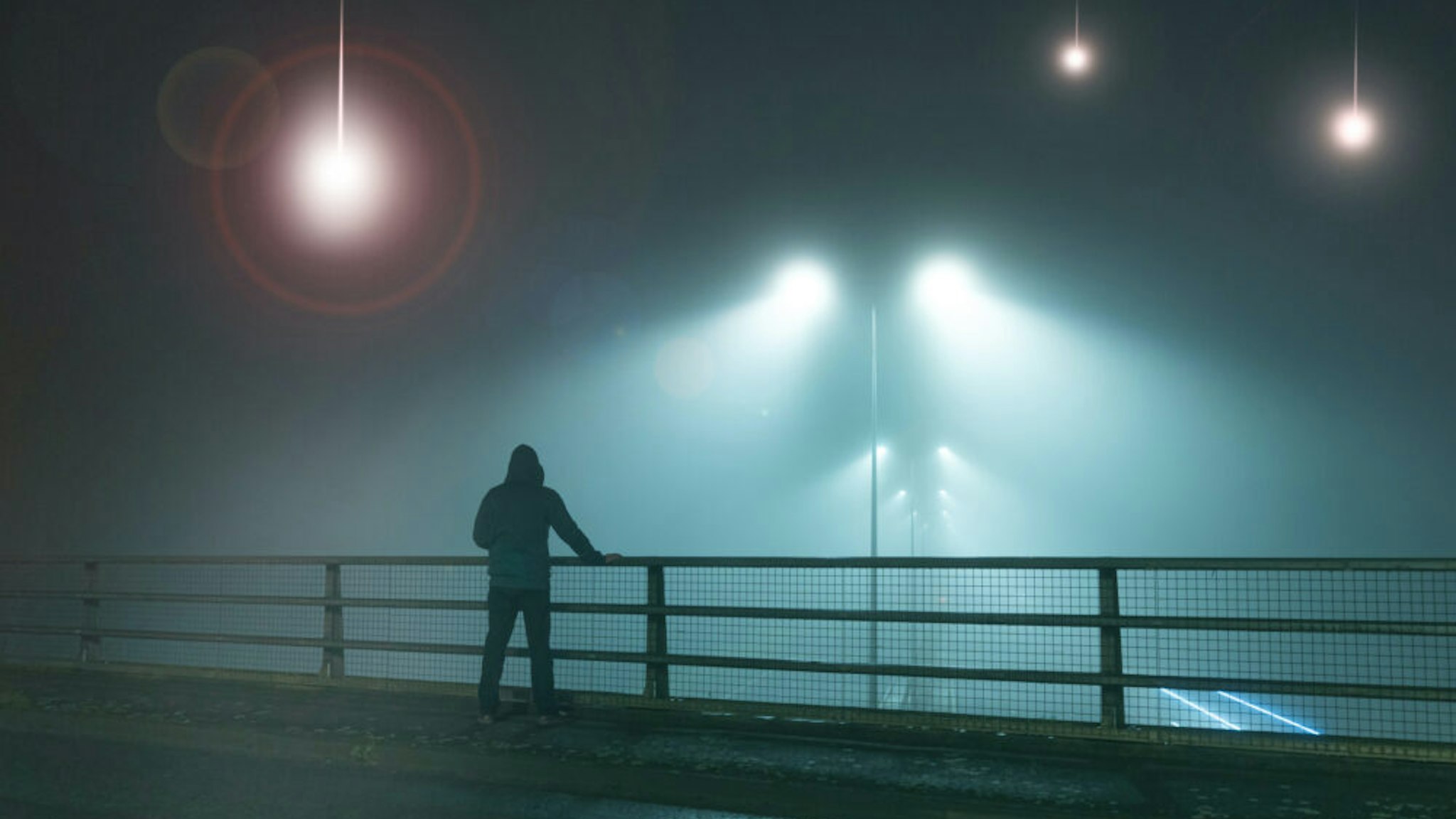 A hooded figure, standing with back to camera on a bridge, looking at UFO alien spaceships coming downfrom the sk., Street lights. On a foggy night.