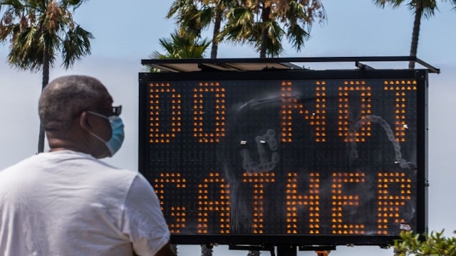 A man wearing a facemask checks his phone near a sign urging people not to gather, while he walks on the beach in Long Beach, California, on July 14, 2020. - California's Governor Gavin Newsom announced a significant rollback of the state's reopening plan on July 13, 2020 as coronavirus cases soared across America's richest and most populous state. (Photo by Apu GOMES / AFP) (Photo by