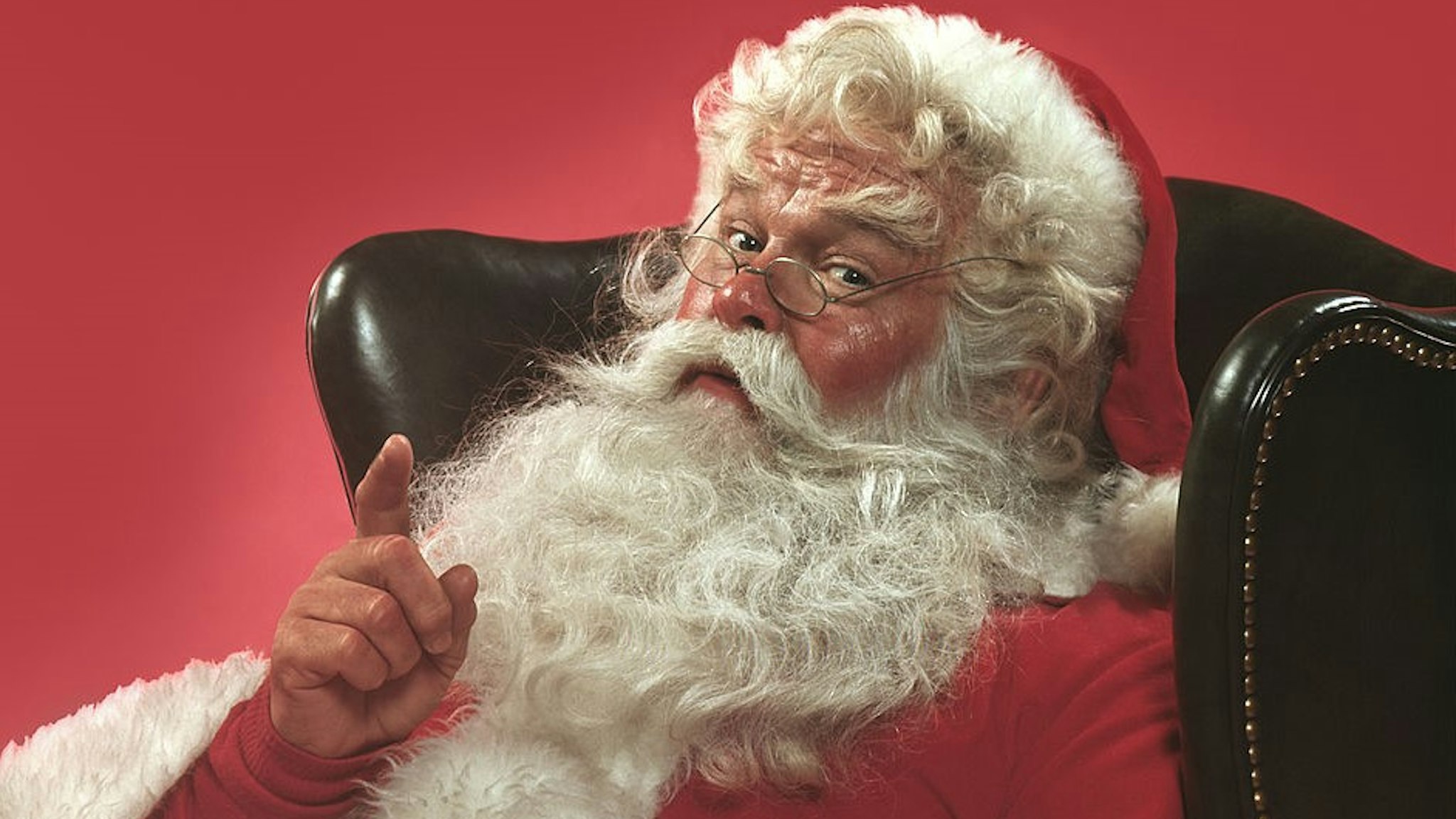 Portrait of Santa Claus sitting in a leather armchair raising one hand in a knowing gesture, 1980. United States. (Photo by T