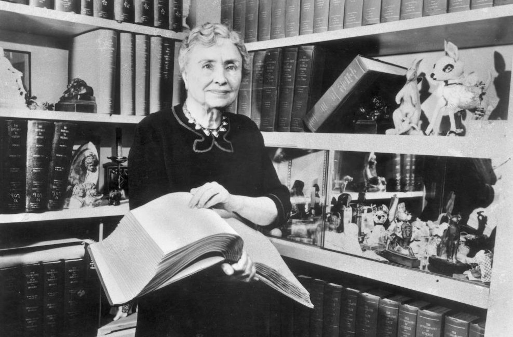 1956: Portrait of American writer, educator and advocate for the disabled Helen Keller (1880 - 1968) holding a Braille volume and surrounded by shelves containing books and decorative figurines. A childhood illness left Keller blind, deaf and mute.