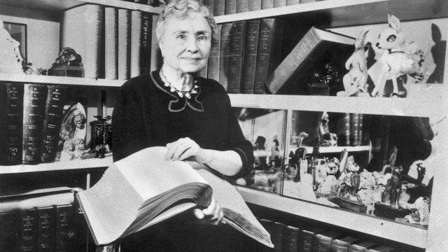 1956: Portrait of American writer, educator and advocate for the disabled Helen Keller (1880 - 1968) holding a Braille volume and surrounded by shelves containing books and decorative figurines. A childhood illness left Keller blind, deaf and mute.