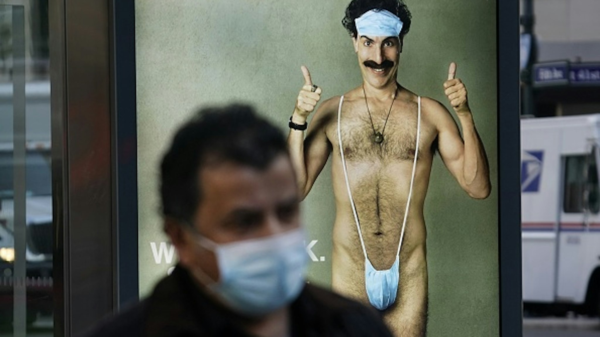A person wearing a mask walks past a bus stop ad on 5th Avenue, October 15, 2020, for the upcoming movie "Borat 2," featuring actor Sacha Baron Cohen, ahead of its release on October 23. - The poster gives reference to the Covid-19 pandemic by replacing Borats iconic green mankini with a face mask.