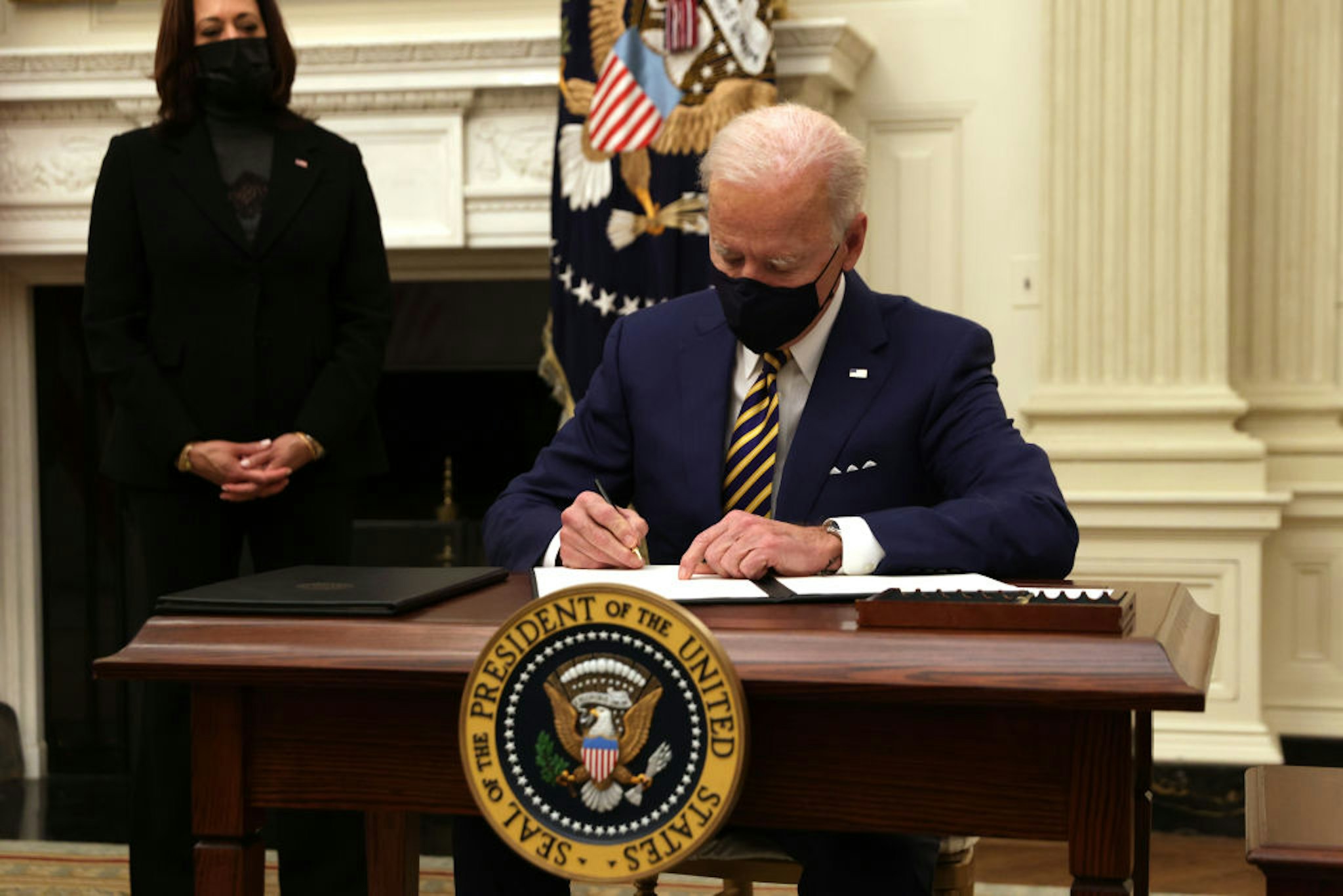 U.S. President Joe Biden signs an executive order as Vice President Kamala Harris looks on during an event on economic crisis in the State Dining Room of the White House January 22, 2021 in Washington, DC.