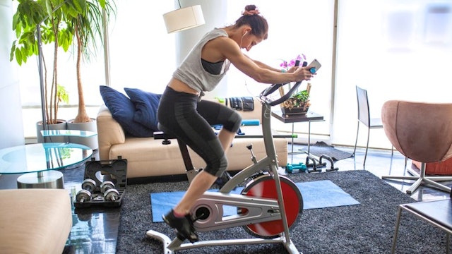 Home Gym under the quarantine, we see a woman using her mobile phone while riding her stationary bike in her apartment, she has conditioned her space in order to keep doing exercise, during the quarantine time of covid19