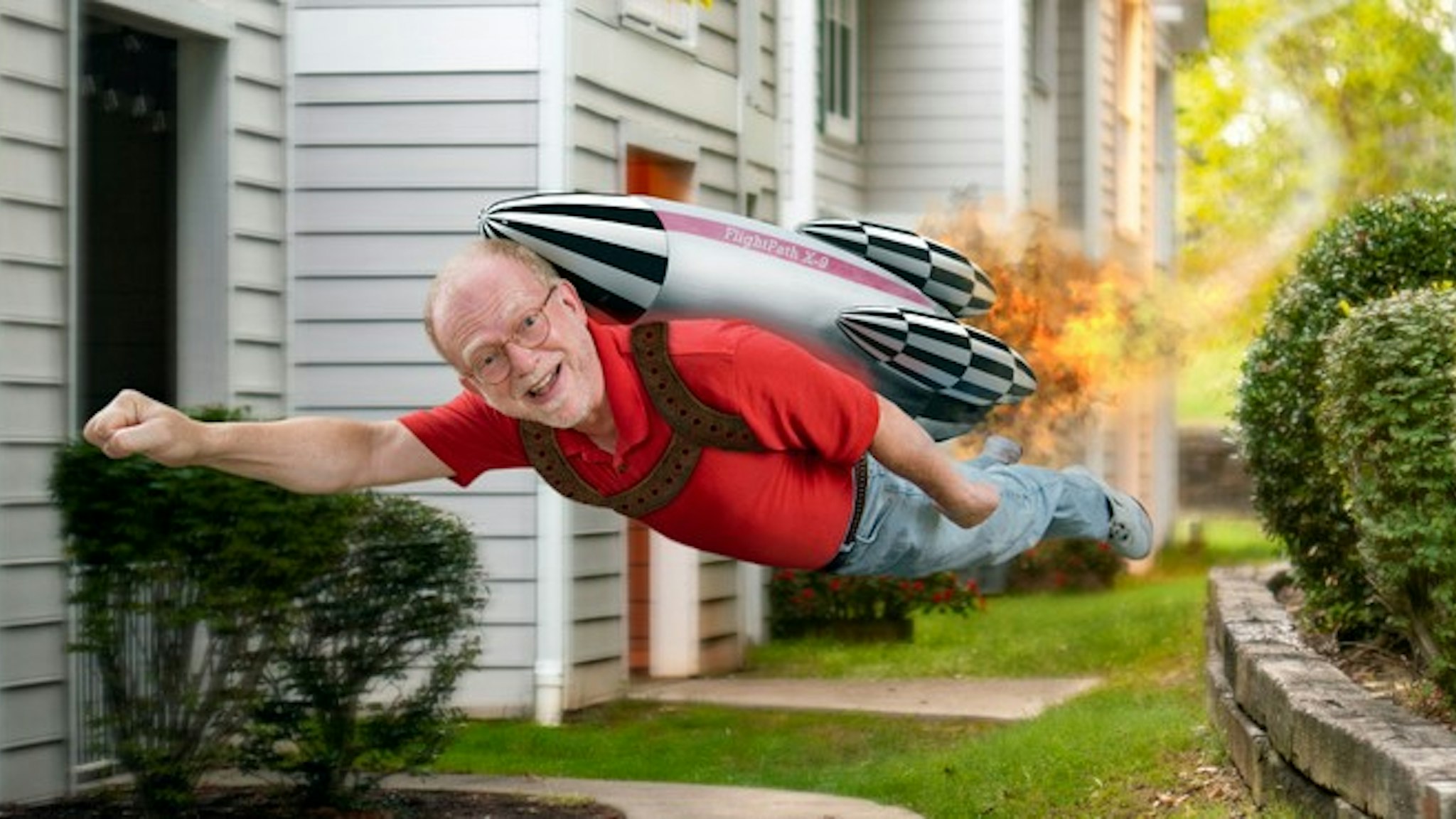 A mature man flies through the air next to an apartment building, a flaming rocket pack is strapped to his back. Smoke marks the trail behind him where he has flown.
