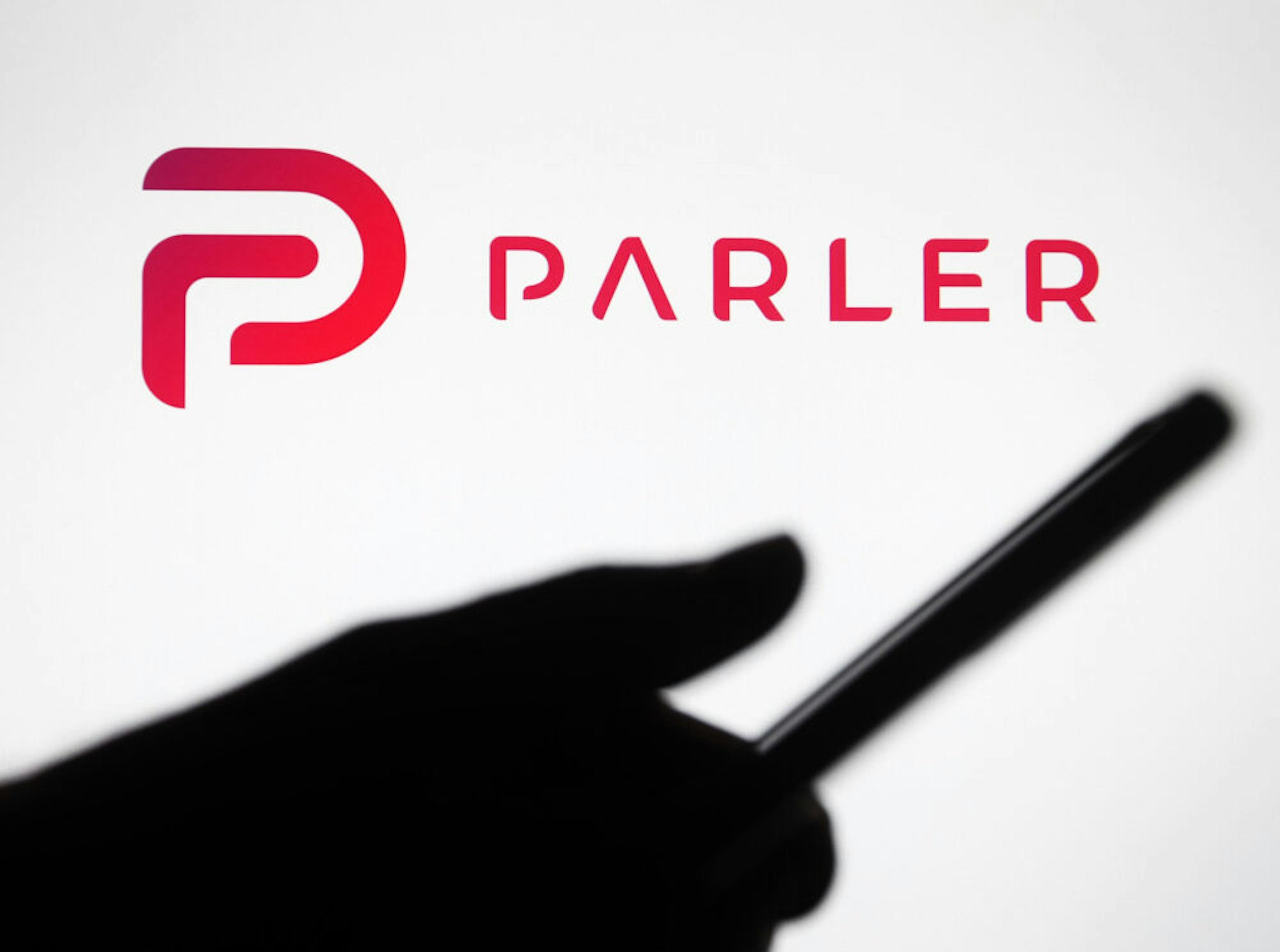 UKRAINE - 2021/01/10: In this photo illustration the Parler logo seen in front of a silhouette hand holding a smartphone. Google, Apple and Amazon have suspended the social networking app Parler. Parler became unavailable in App Store, Google Play and Amazon Web Services, reportedly as said insufficient control over user posts that encouraged violence, reportedly by media.