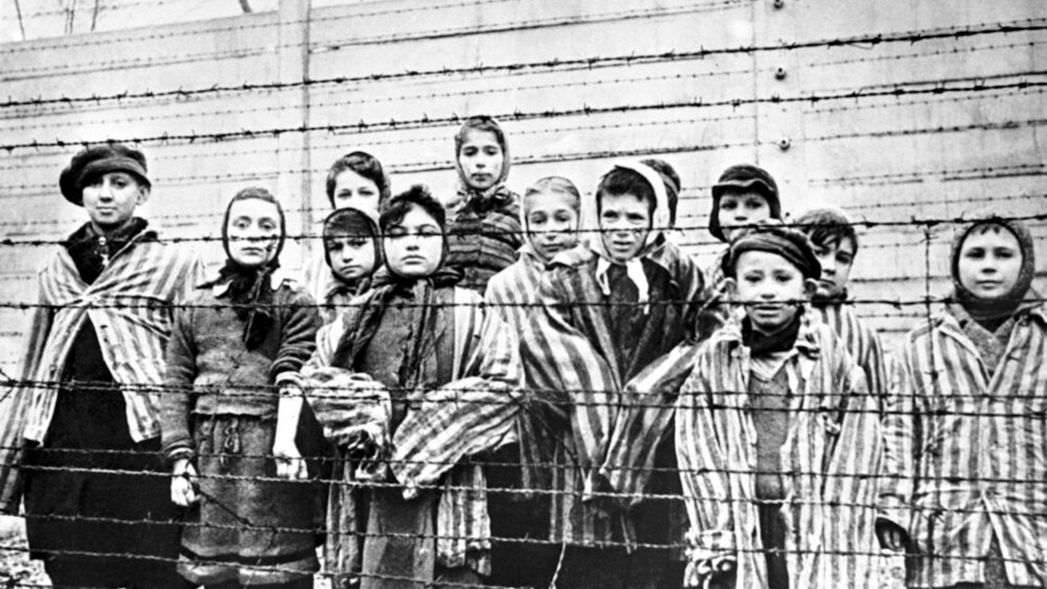 A group of child survivors behind a barbed wire fence at the Nazi concentration camp at Auschwitz-Birkenau in southern Poland, on the day of the camp’s liberation by the Red Army, 27th January 1945. Photo taken by Red Army photographer Captain Alexander Vorontsov during the making of a film about the liberation of the camp. The children were dressed in adult uniforms by the Russians. The children are (left to right): Tomy Schwarz (later Shacham), Miriam Ziegler, Paula Lebovics (front), Ruth Webber, Berta Weinhaber (later Bracha Katz), Erika Winter (later Dohan), Marta Weiss (later Wise), Eva Weiss (later Slonim), Gabor Hirsch (just visible behind Eva Weiss), Gabriel Neumann, Robert Schlesinger (later Shmuel Schelach), Eva Mozes Kor, and Miriam Mozes Zeiger.