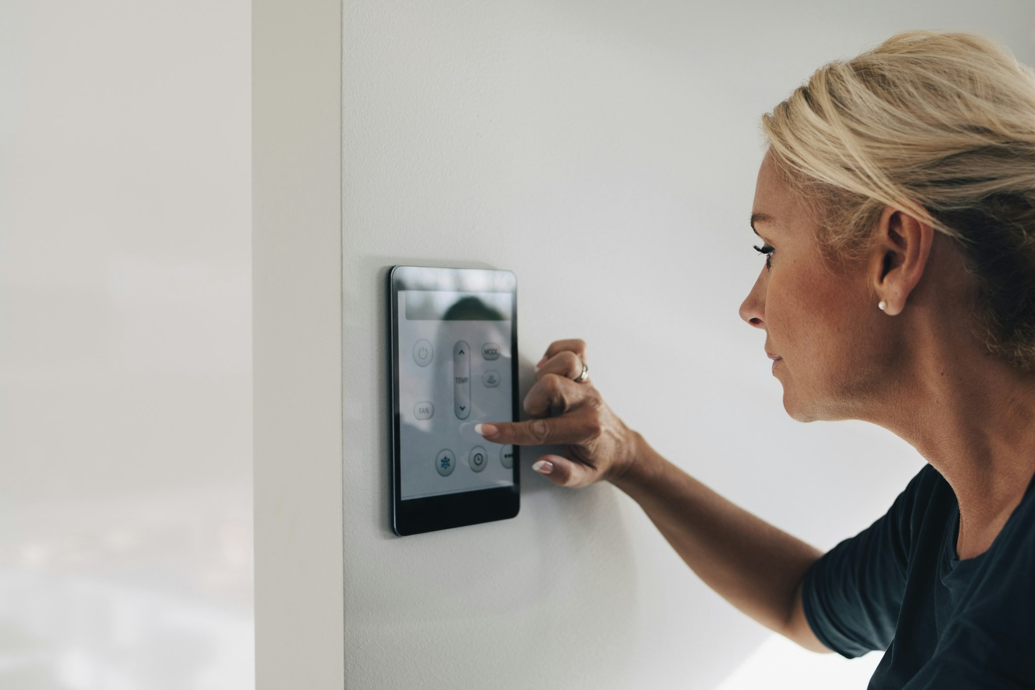 Blond woman adjusting thermostat using digital tablet mounted on white wall at home - stock photo