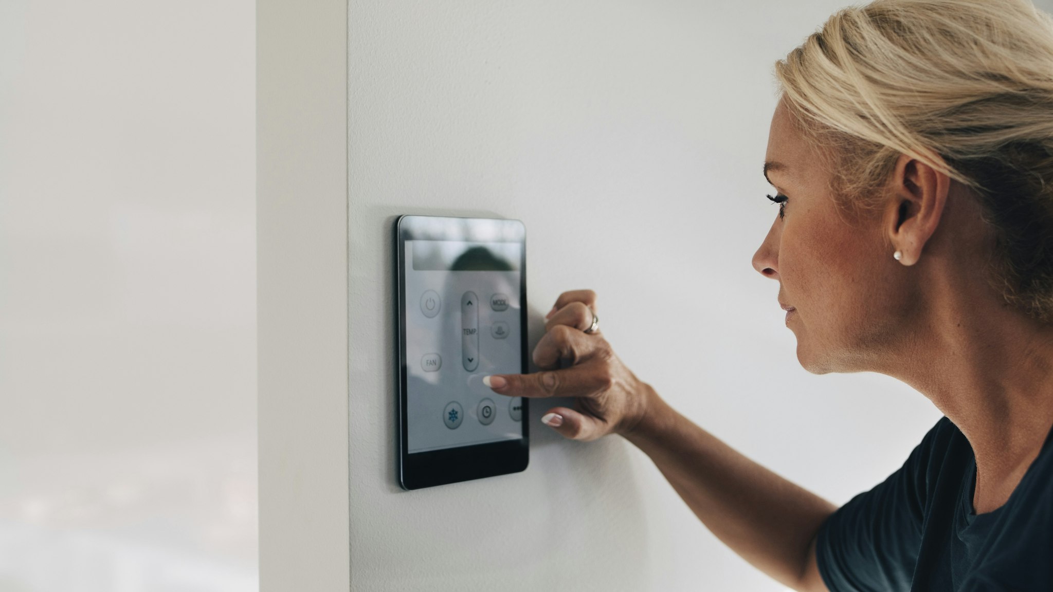 Blond woman adjusting thermostat using digital tablet mounted on white wall at home - stock photo